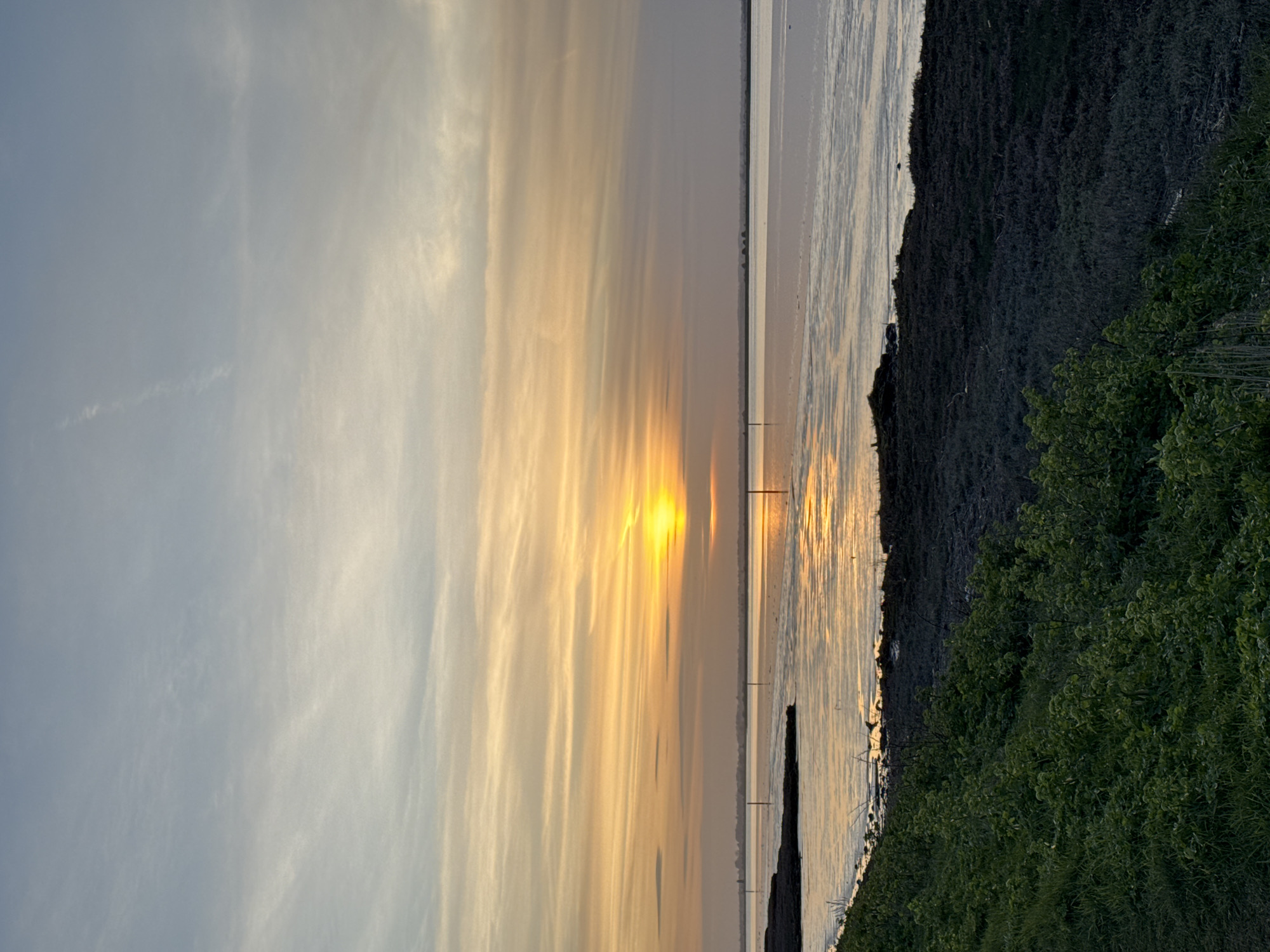 The image shows a sunset over a body of water, likely Breydon Water near Great Yarmouth, United Kingdom. The sun is near the horizon, casting a bright golden light across the sky and reflecting in the water. The sky shows wispy clouds. The foreground includes a shoreline with vegetation, and in the distance, poles are visible in the water. No people are visible in the scene.