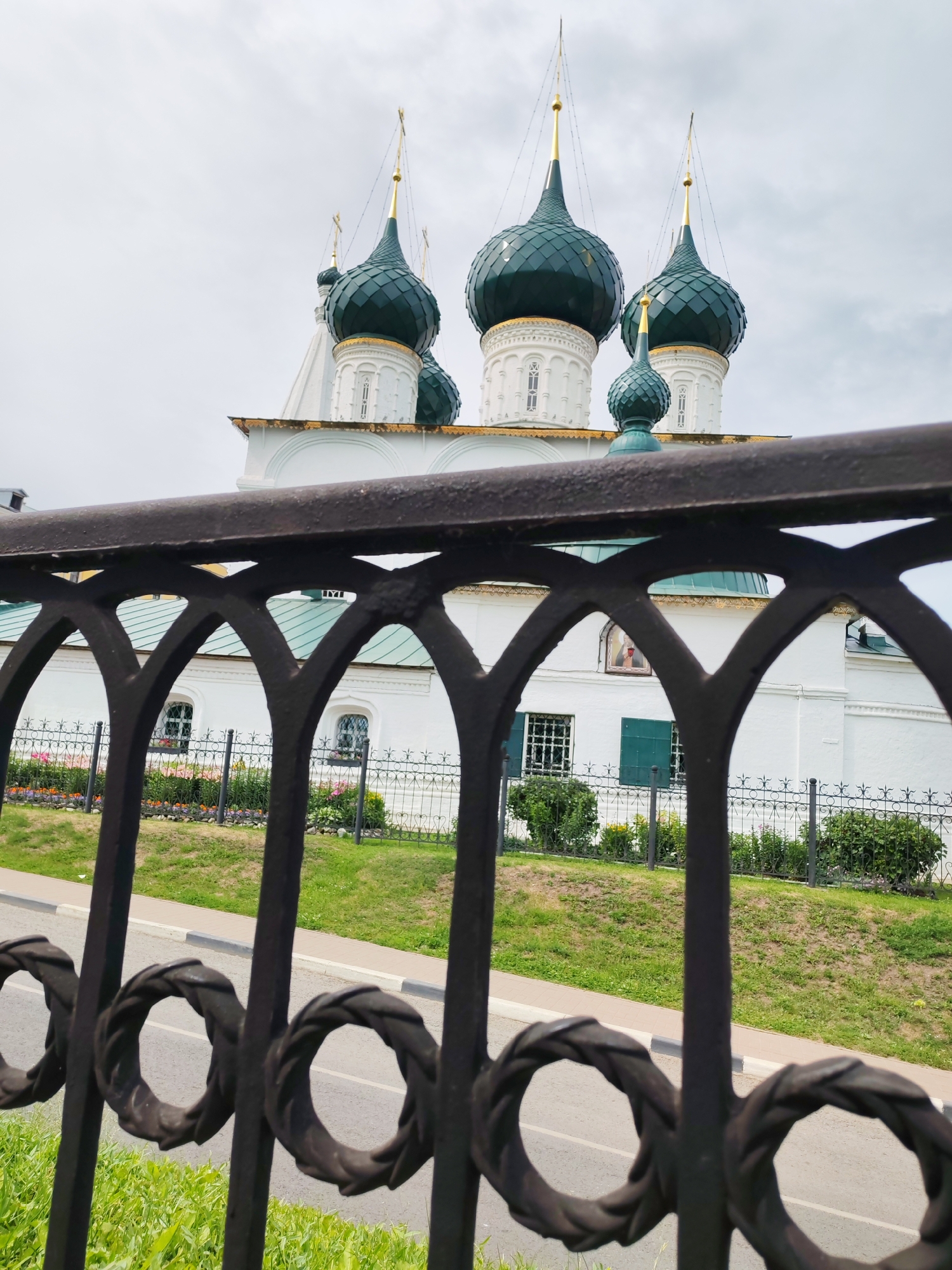 An ornate black metal fence is in the foreground, with a building with several green domes and gold spires in the background. The building appears to be a church, possibly Orthodox, given the style of the domes. The church is white and has arched windows. There is a small garden with flowers and greenery in front of the building, separated from the street by a decorative black metal fence. The street is paved and bordered by a sidewalk and a grassy area. The sky is overcast. The location is identified as Rusanovo, Russia. There are no people visible in the image.