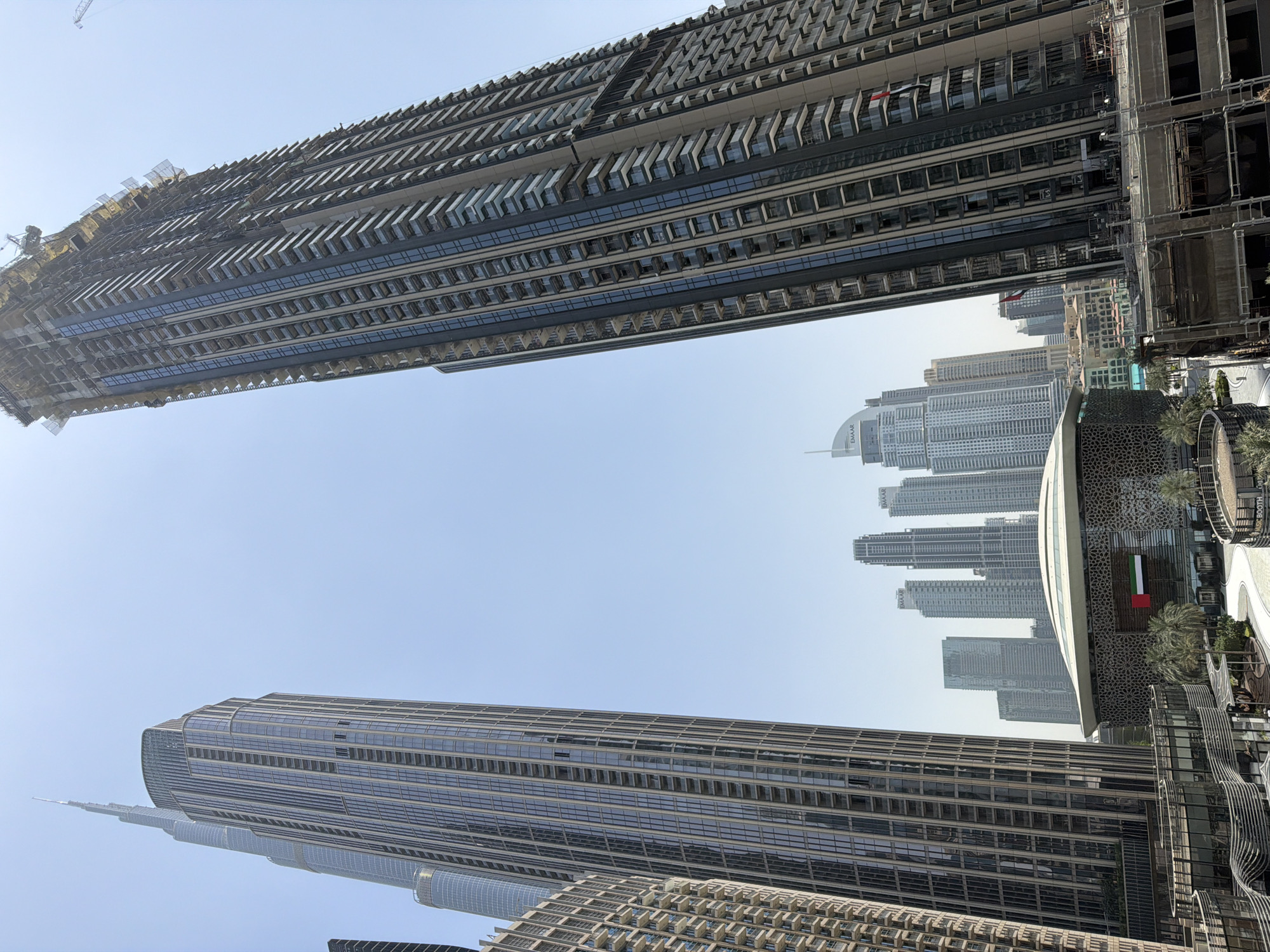 The image captures a low-angle view of the urban landscape in Dubai, United Arab Emirates. Two skyscrapers frame the composition, with one under construction on the top and the other completed on the bottom. A crane is visible on the top building. Other buildings are in the background. A building with a flag is visible on the right of the image. The sky is a clear blue, indicating a sunny day. There are no people visible in the frame.