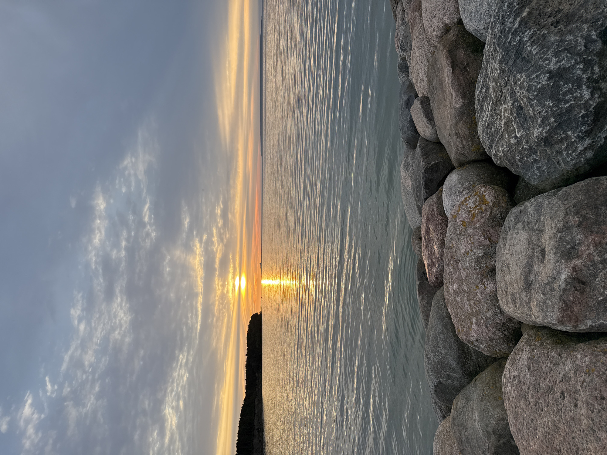 The image shows a sunset over a body of water, likely the Baltic Sea, with a line of trees on the left horizon and a rocky shore in the foreground. The sun is low in the sky, casting a golden reflection on the rippling water. Clouds are present in the sky, tinged with orange and yellow hues from the setting sun. The rocks in the foreground are large and varied in color and texture. There is a ship visible in the distance on the water. This scene could be a view from the coast of Tallinn, Estonia, a city known for its seaside location and historical significance.