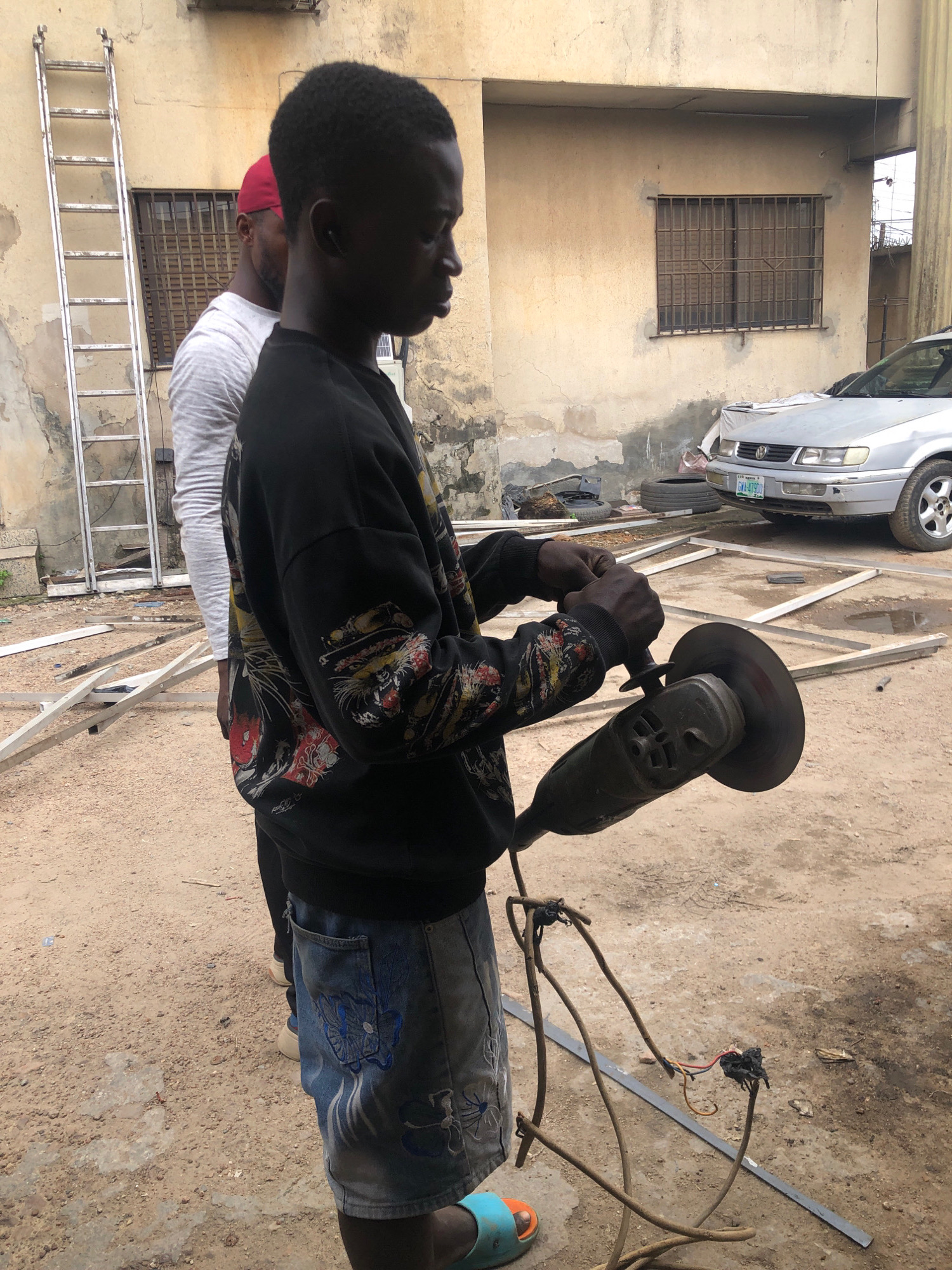 In a dusty, outdoor setting in Ikeja, Nigeria, two young men are engaged in what appears to be construction or fabrication work. The foreground is dominated by a young man wearing a black sweatshirt with a colorful, graphic print and denim shorts adorned with embroidered blue flowers. He is actively using a handheld grinding tool, holding it with both hands as sparks fly from the spinning disc. The tool is connected to a power source by a bundle of wires that are taped together. To the left and slightly behind him, another young man, wearing a red cap and a grey long-sleeved shirt, stands partially obscured. In the background, a pale yellow building with barred windows is visible. A tall, silver metal ladder leans against the building. Several pieces of metal framing are laid out on the ground, suggesting ongoing construction. A silver Volkswagen car is parked in the background, and nearby, two tires are stacked. The ground is covered in loose dirt and small stones, with some puddles of water indicating recent rain. The overall scene depicts an active and somewhat rugged work environment within an urban Nigerian context.