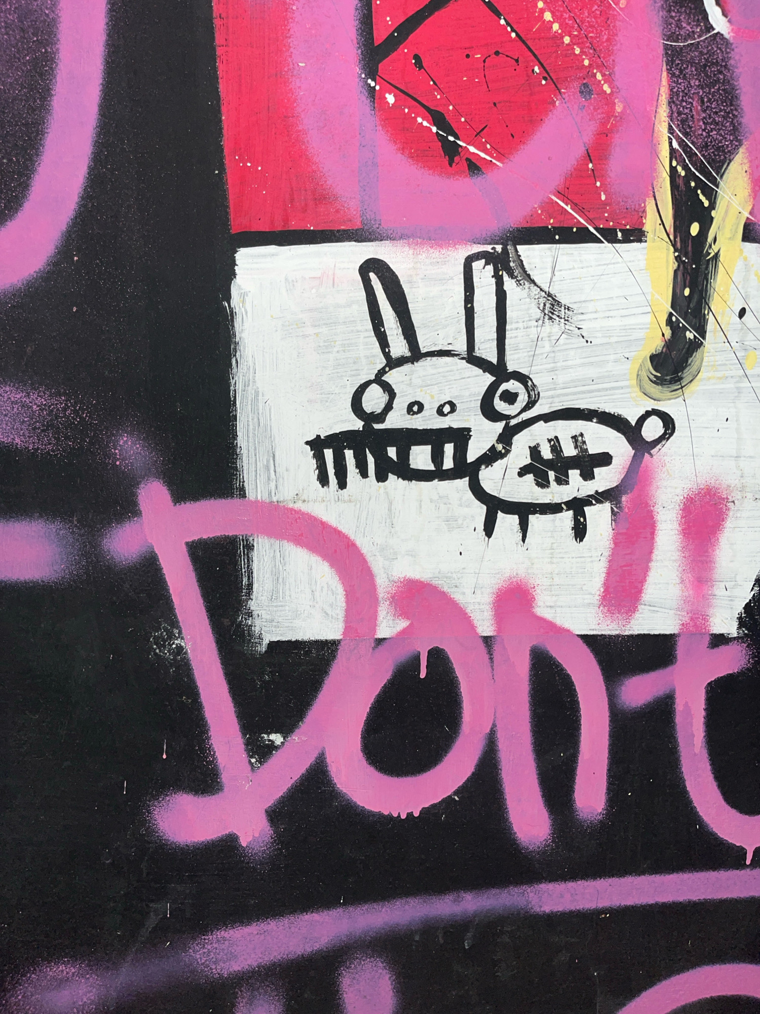 This is a close-up, high-angle photograph of graffiti art on a black wall, with elements of pink, white, and red. The graffiti includes a crudely drawn cartoon rabbit with wide eyes and a stylized body, seemingly placed on a white background within the black wall. Below the rabbit, large pink bubble letters spell out "Don't", with drips of pink paint visible. A thinner, lighter purple spray-painted line runs horizontally below the "Don't" graffiti. The overall style is energetic and abstract, typical of street art. The photograph was taken in the city of Surakarta, Indonesia.