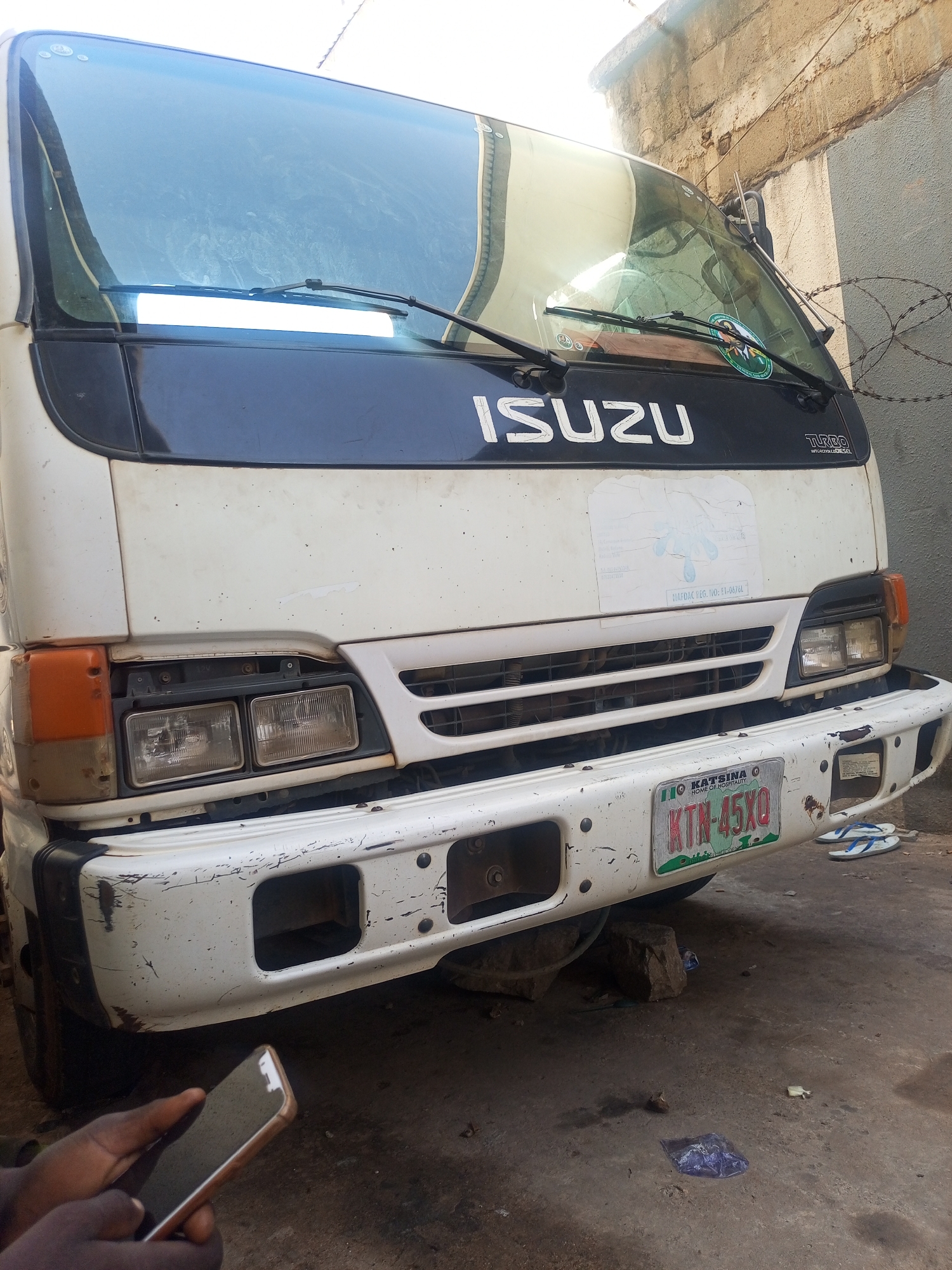 The image features the front of a white Isuzu truck with some damage to its bumper and body. The license plate is visible, reading "KTN-45XQ" and marked with "Katsina, Home of Hospitality," indicating it is from Katsina State, Nigeria. The background shows a building with barbed wire. A person's hand holding a smartphone is visible in the bottom left corner of the frame. The word, "Turbo Intercooler" appears on the upper right of the front of the truck. The vehicle appears to be undergoing repair based on an object under the bumper. The location is possibly Maiburiji, Nigeria, as the person taking the photo is there.