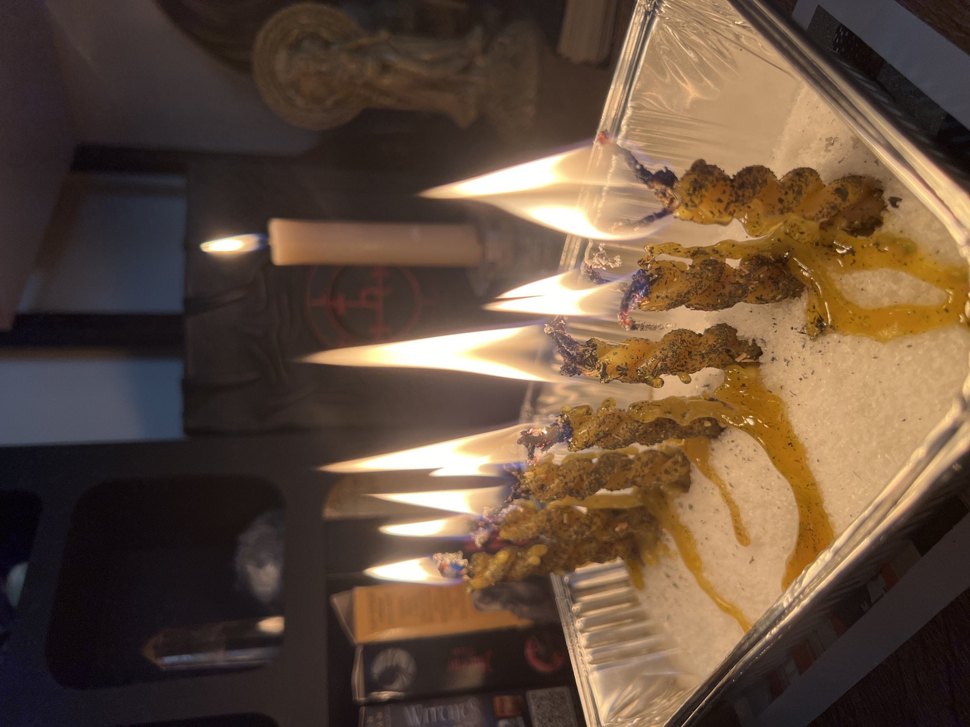 The image depicts a close-up view of seven lit candles arranged in a rectangular foil pan filled with white granular material, possibly salt or sand. The candles are thin and twisted, with a rough, textured surface, and appear to be melting, with golden wax dripping onto the white material. The flames are bright and flickering, casting a warm glow. In the background, shelves are visible, containing various objects including books, crystals, and a statue. The overall scene suggests an indoor setting, possibly a room with shelves displaying decorative or ritualistic items. The image does not provide any information that would allow for determination of a specific location in the city of Tychy, Poland.