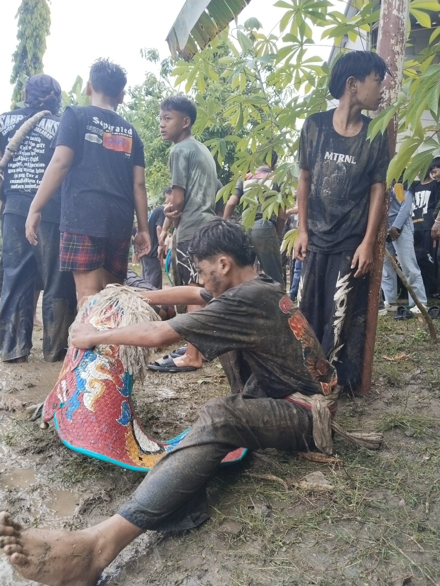 The image shows a group of people in what appears to be a muddy outdoor setting, possibly in Karang Tengah, Indonesia. Several young men are visible. One is kneeling in the mud, interacting with a colorful, woven object that seems to be part of a traditional cultural item.  The individual is covered in mud from head to toe. He is holding the object with both hands. Other people are standing around, some wearing t-shirts with English writing. The surroundings consist of what appears to be a yard with grass and muddy patches. Some tropical foliage and trees are visible in the background.