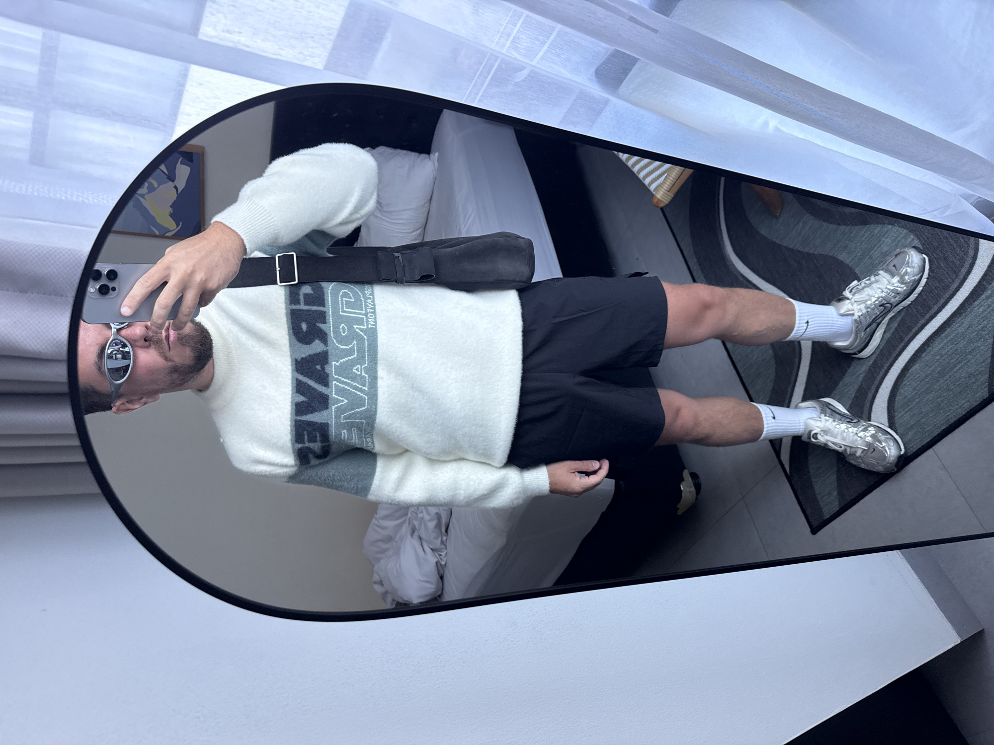 A man stands in front of an oval mirror, holding a phone up to take a picture of himself in North Kuta, Indonesia. He is wearing sunglasses, a cream and gray striped sweater, black shorts, white socks, and silver sneakers. A black messenger bag hangs across his torso. The background visible in the mirror includes a bed with white linens, a rug with gray and black swirls, and a window covered with white curtains. The mirror is propped up against a white wall.