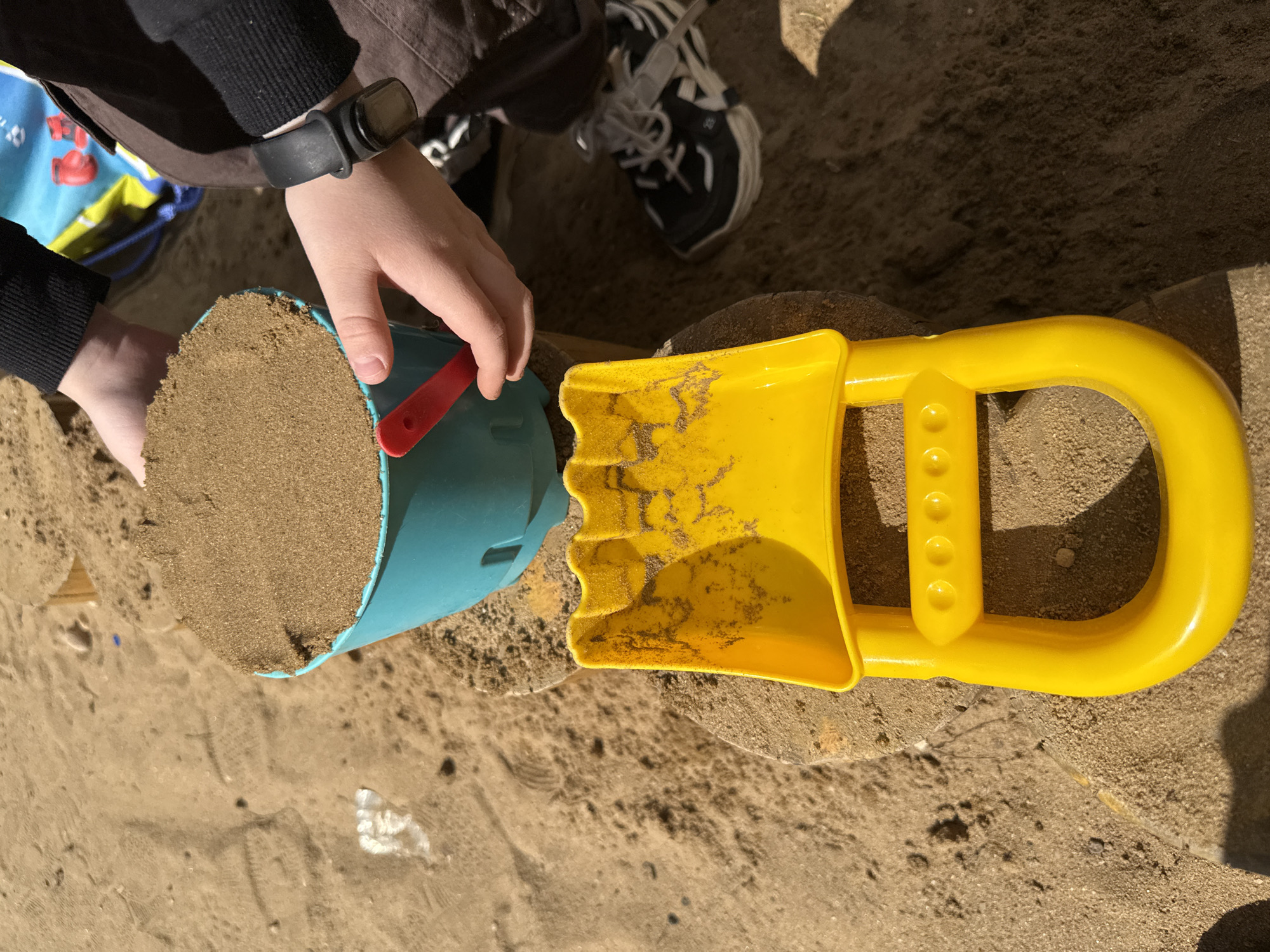 A child is using a blue bucket filled with sand to fill a yellow plastic sand shovel in a sand pit. The child is wearing a black watch on their left wrist, a black long-sleeved shirt, and brown pants. The child's shoes are black and white. A blue bag with red and yellow designs is partially visible in the background. The environment is a sandy area, with varying textures and slight discolorations. No evidence suggests that the image was taken in Lyubertsy, Russia.