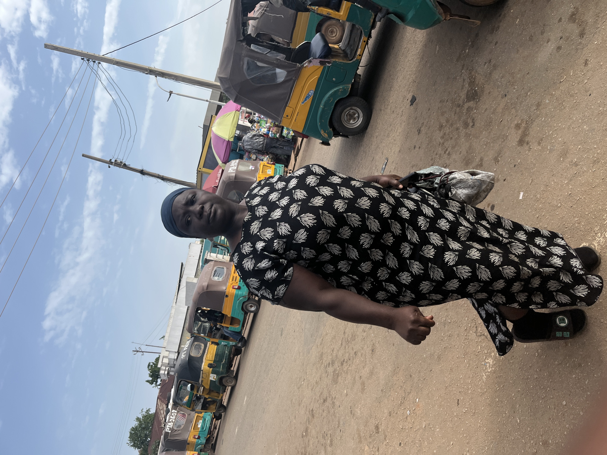 An African woman wearing a black and white patterned dress and a dark headscarf stands on a dirt road in Gura Top, Nigeria, facing the camera. She holds a grey bag in her left hand. Behind her, several brightly colored auto-rickshaws, common modes of transportation in Nigeria, are parked in a line along the road. The sky above is partly cloudy. A few market stalls are visible in the background, with people shopping. Power lines stretch across the sky, supported by wooden poles.