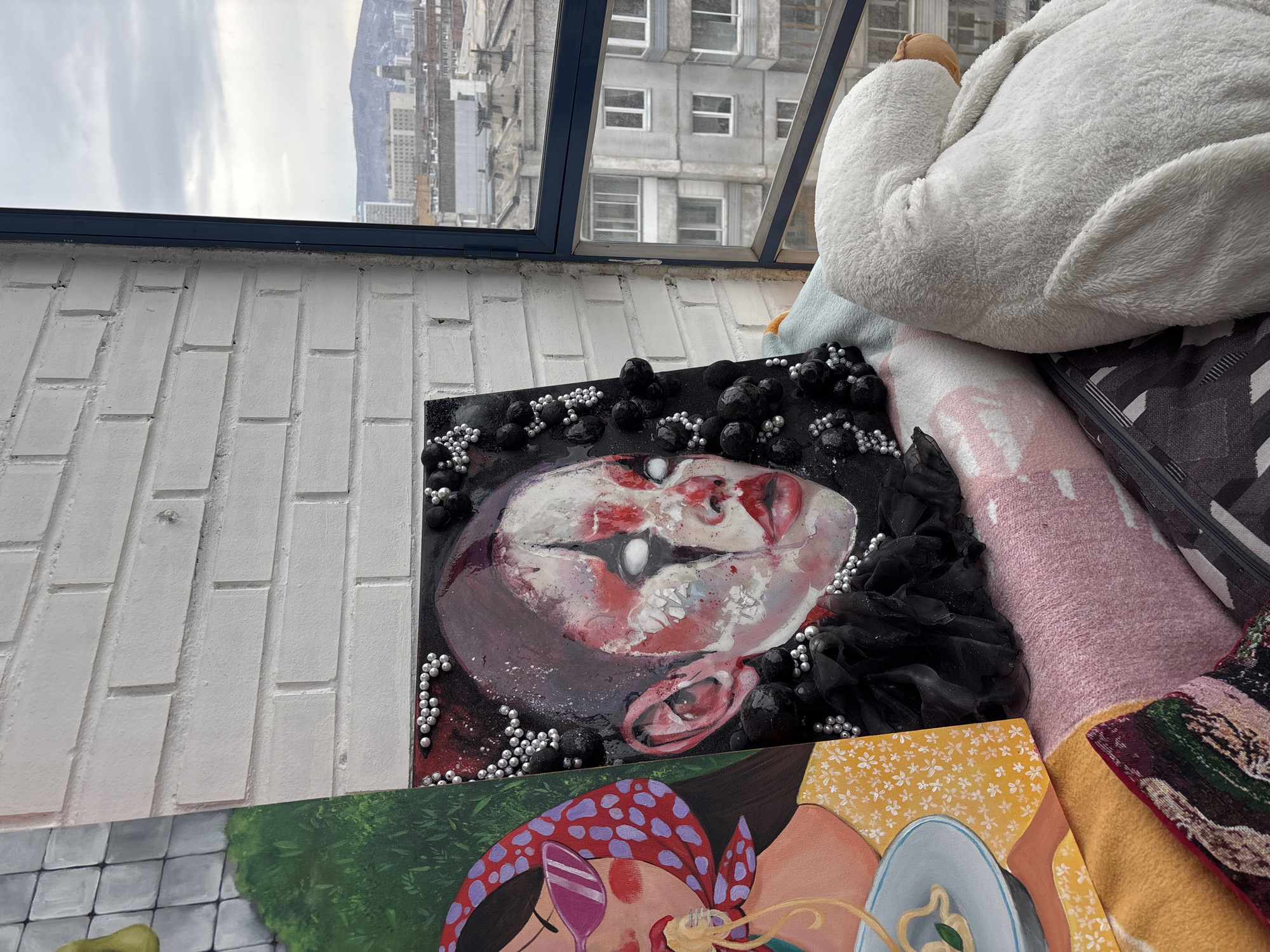An indoor scene displays two artworks positioned near a window, offering an urban panorama of Krasnoyarsk, Russia. On the left, a partial view of a vertical canvas depicts a light-skinned woman wearing purple sunglasses and a red polka-dotted headscarf, eating yellow spaghetti from a light blue bowl. Adjacent to it, centrally positioned, is a rectangular, mixed-media artwork. This piece features a stylized face with pale skin, prominent red and black facial markings resembling clown makeup, and opaque white eyes. The artwork is textured with clusters of small, shiny silver spherical beads, larger, rough-textured black spheres, and ruffled dark fabric elements around the lower part of the face.
Both artworks are supported by a stack of folded textiles, which include sections of pink, orange, and patterned fabrics. To the right, a large, white, fluffy plush toy, resembling a goose or swan, is visible, positioned near the window with its head tucked.
The background shows a dark-framed window looking out onto an urban landscape under an overcast sky. This view encompasses multiple multi-story grey residential and commercial buildings, industrial-style roofs, and in the far distance, snow-dusted hills or mountains consistent with the topography surrounding Krasnoyarsk. A white brick wall forms the interior backdrop to the left of the window.