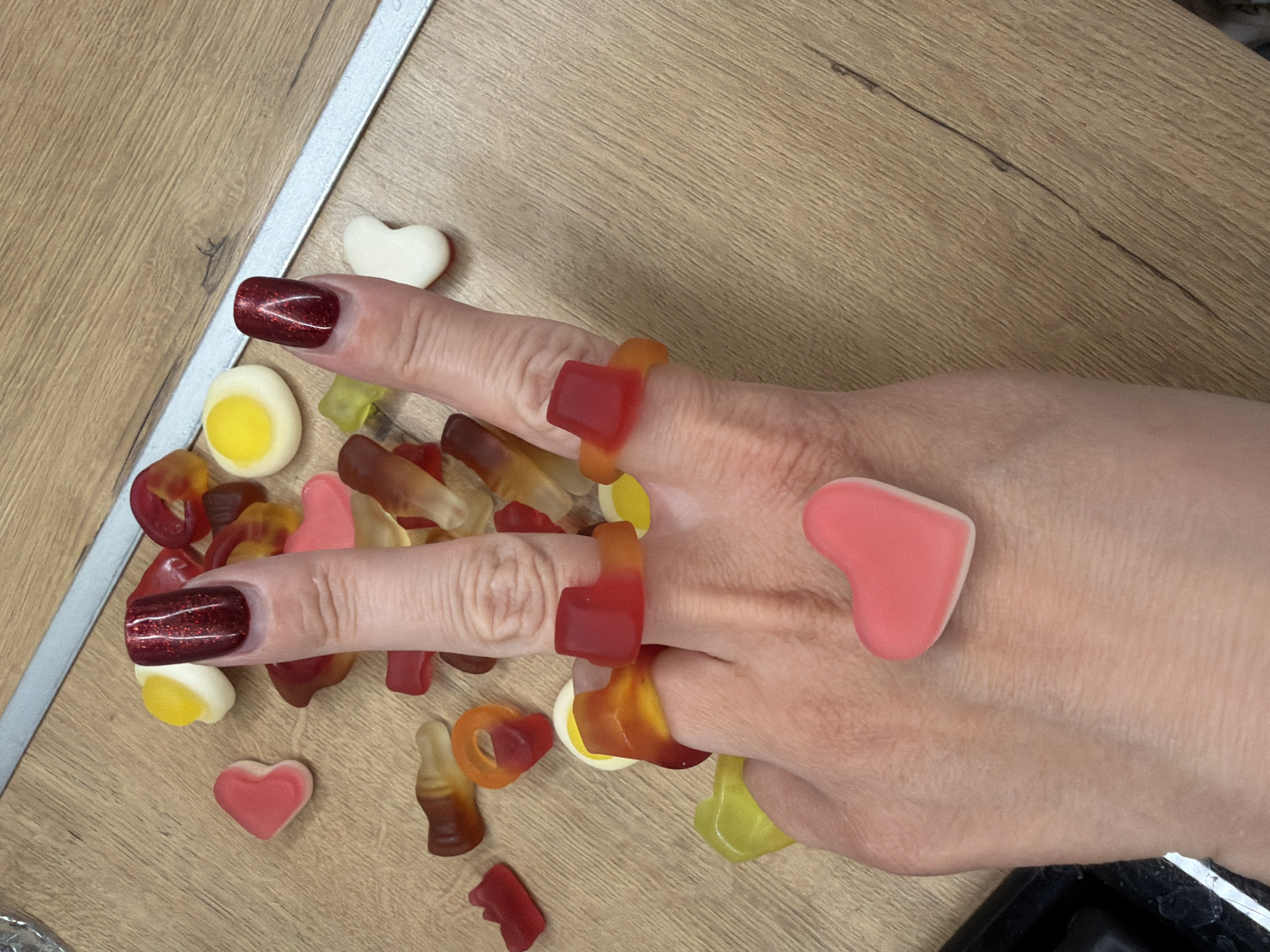 A hand wearing gummy candy rings and adorned with gummy candy hearts is displayed on a light brown, wood-grain surface. The fingernails of the hand are painted a dark red with glitter. Scattered around the hand are assorted gummy candies, including eggs, bottles, and rings. The background features a light gray trim along the wood-grain surface. No information suggests the image was taken in Votkinsk, Russia.