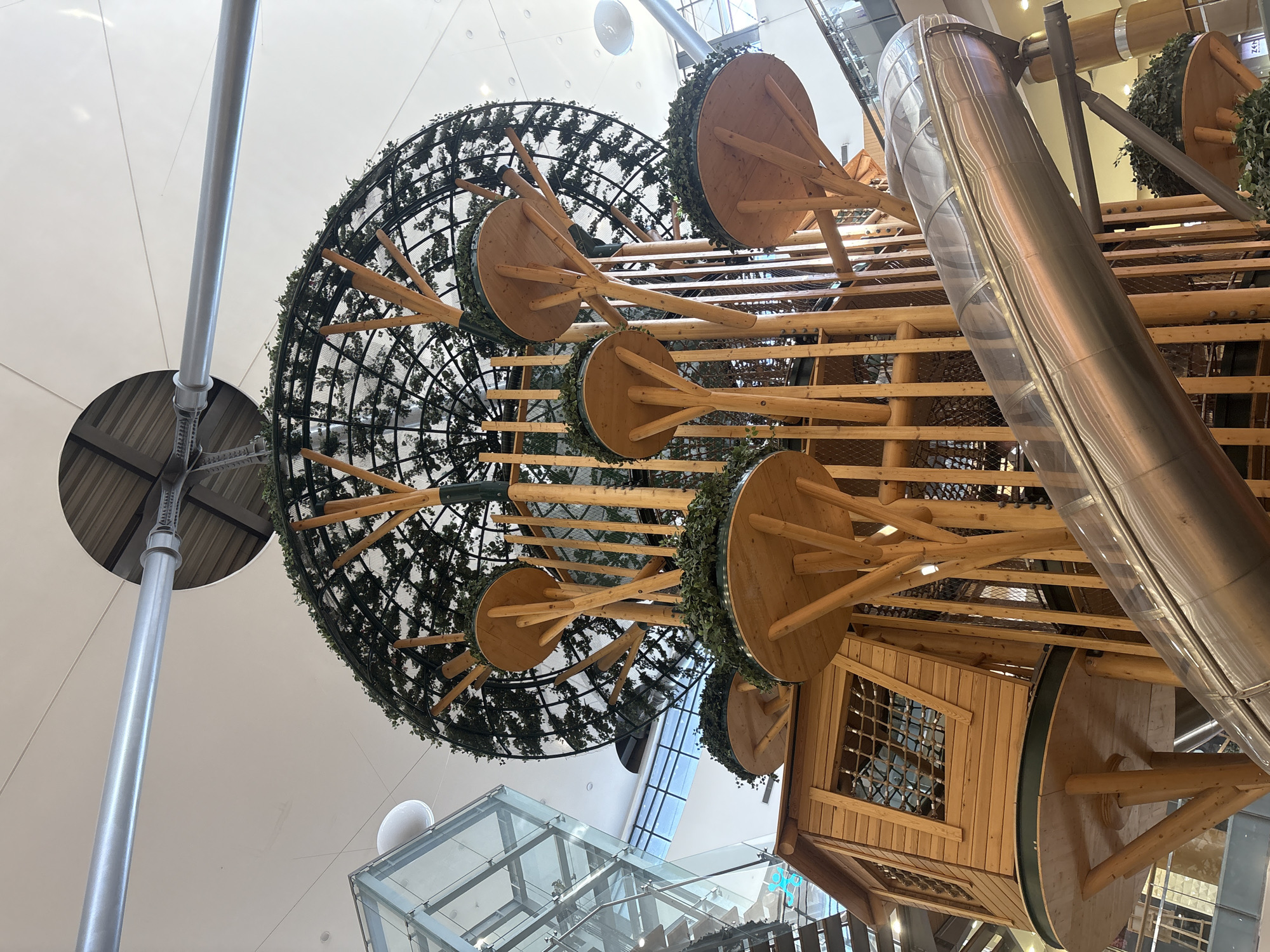 An indoor children's playground structure, resembling a large tree with multiple levels, is the primary subject of the image. The structure is made of wood and metal, with circular platforms and a wooden playhouse. A large, transparent tube slide spirals down from one of the upper levels. The playground is set against a backdrop of a modern building interior, with a high ceiling, large windows, and escalators visible in the distance. The lighting suggests an indoor environment, possibly a shopping mall or entertainment center. There are no people visible in the image.