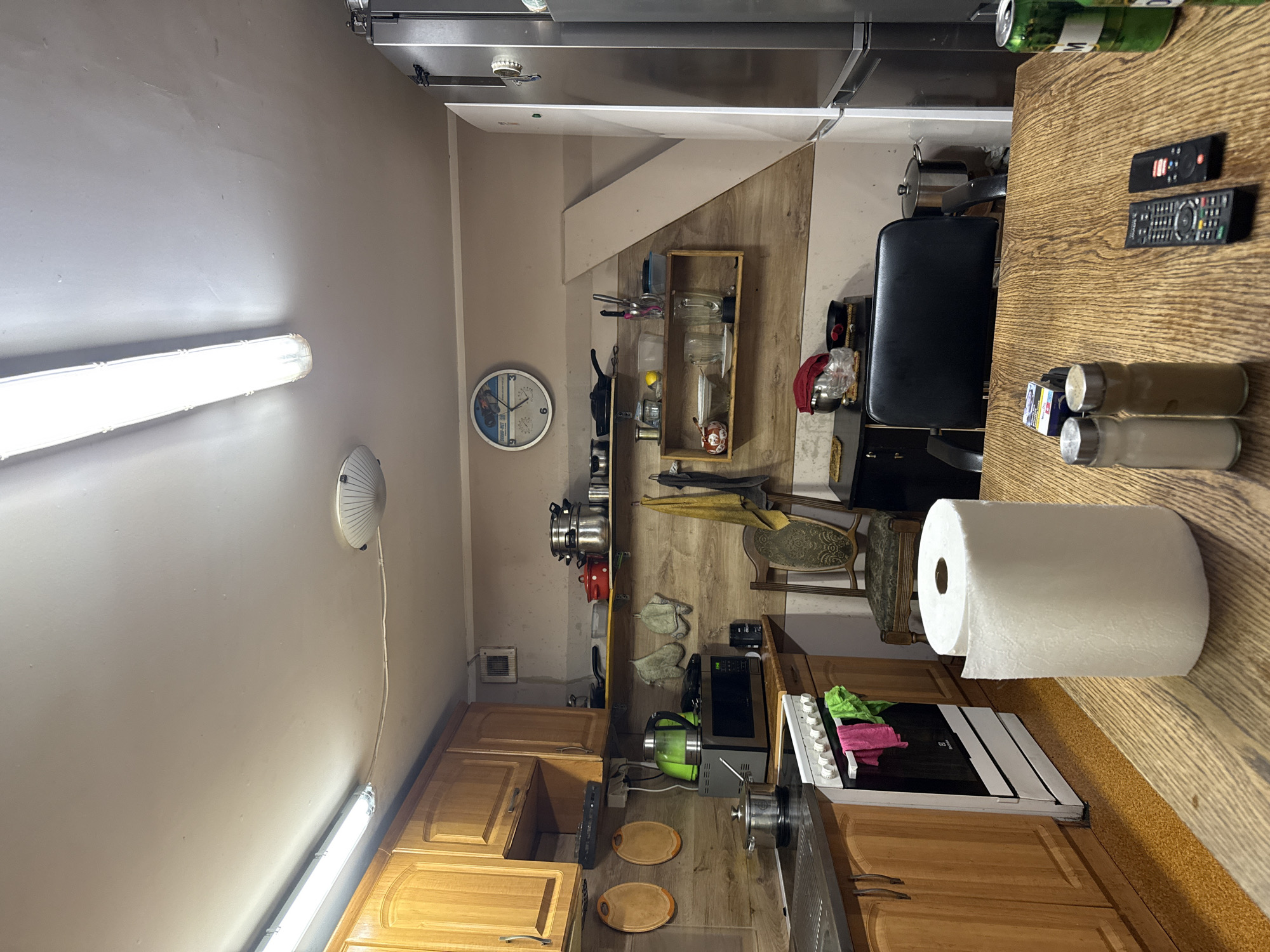 The image depicts a rustic kitchen in Wąwelnica, Poland. The kitchen is equipped with wooden cabinets, a white oven with colorful towels draped over it, and a microwave. Above the counter, shelves hold various kitchenware, including pots, pans, and a blender. A round clock hangs on the wall, and a tall refrigerator stands to the right. In the foreground, a wooden table is set with a roll of paper towels, two shakers, and two remote controls. A can of beer is also visible on the table. The lighting appears to be from fluorescent ceiling fixtures. The overall impression is one of a lived-in, functional kitchen.