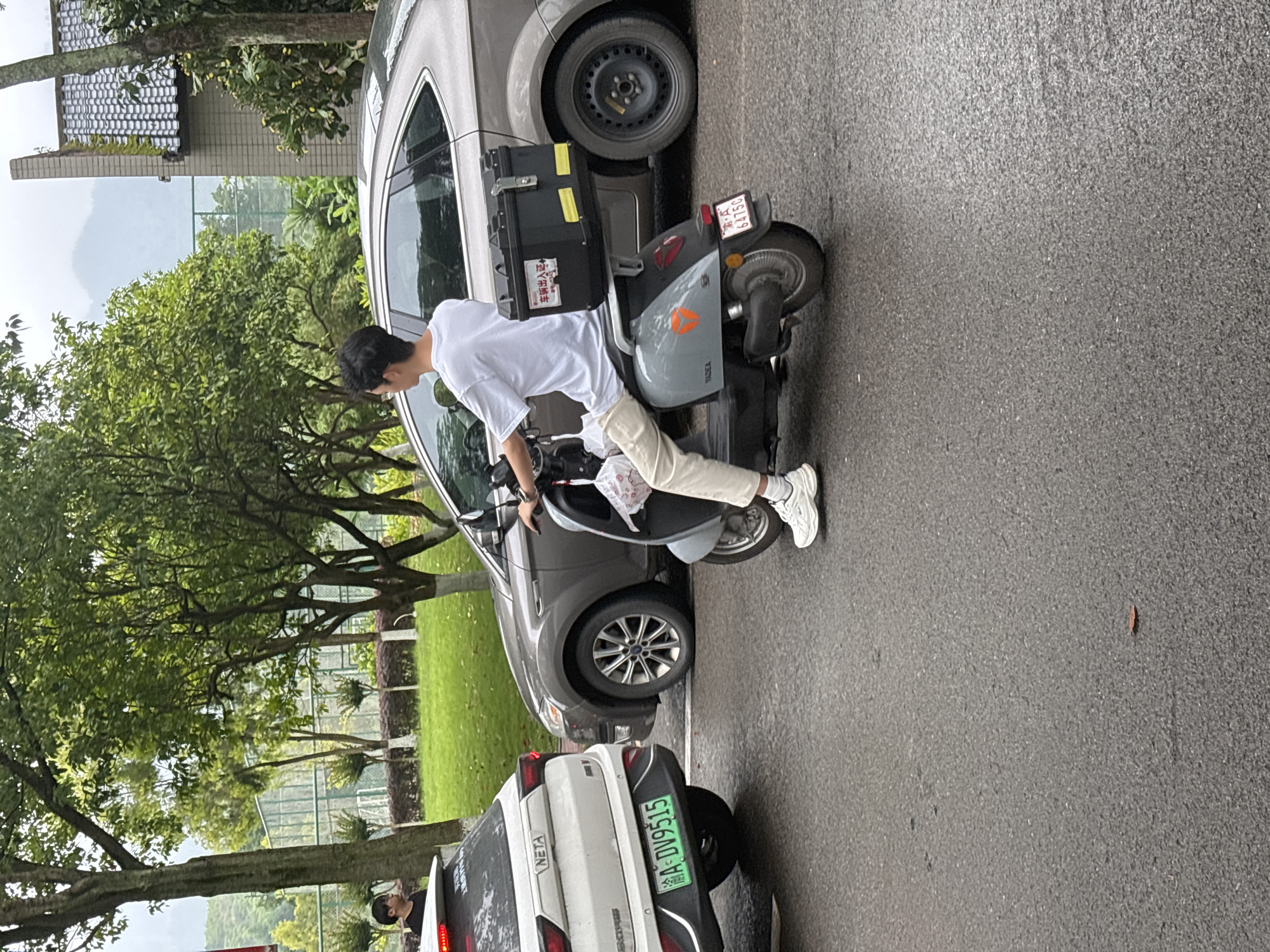 A person is sitting on a gray scooter parked on a paved surface in Jiangjin, China. The scooter is parked between a gray car and a white car. The license plate on the white car reads "A-DV9515." There are trees and a green lawn visible in the background. The weather appears overcast.
