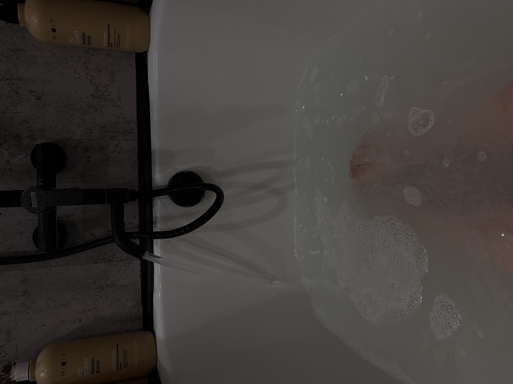 The image shows a bathtub being filled with water. A black faucet with a curved spout is mounted on the side of the tub. Two bottles of "RE: START Keratin Bomb" hair products from MINIT, one shampoo and one conditioner, sit on the ledge of the tub. Bubbles are visible in the water, suggesting soap or bath product has been added. A foot is partially visible in the water. The photo appears to have been taken from an overhead perspective. The location details (Reshetnikovo, Russia) are not discernible from the image content.