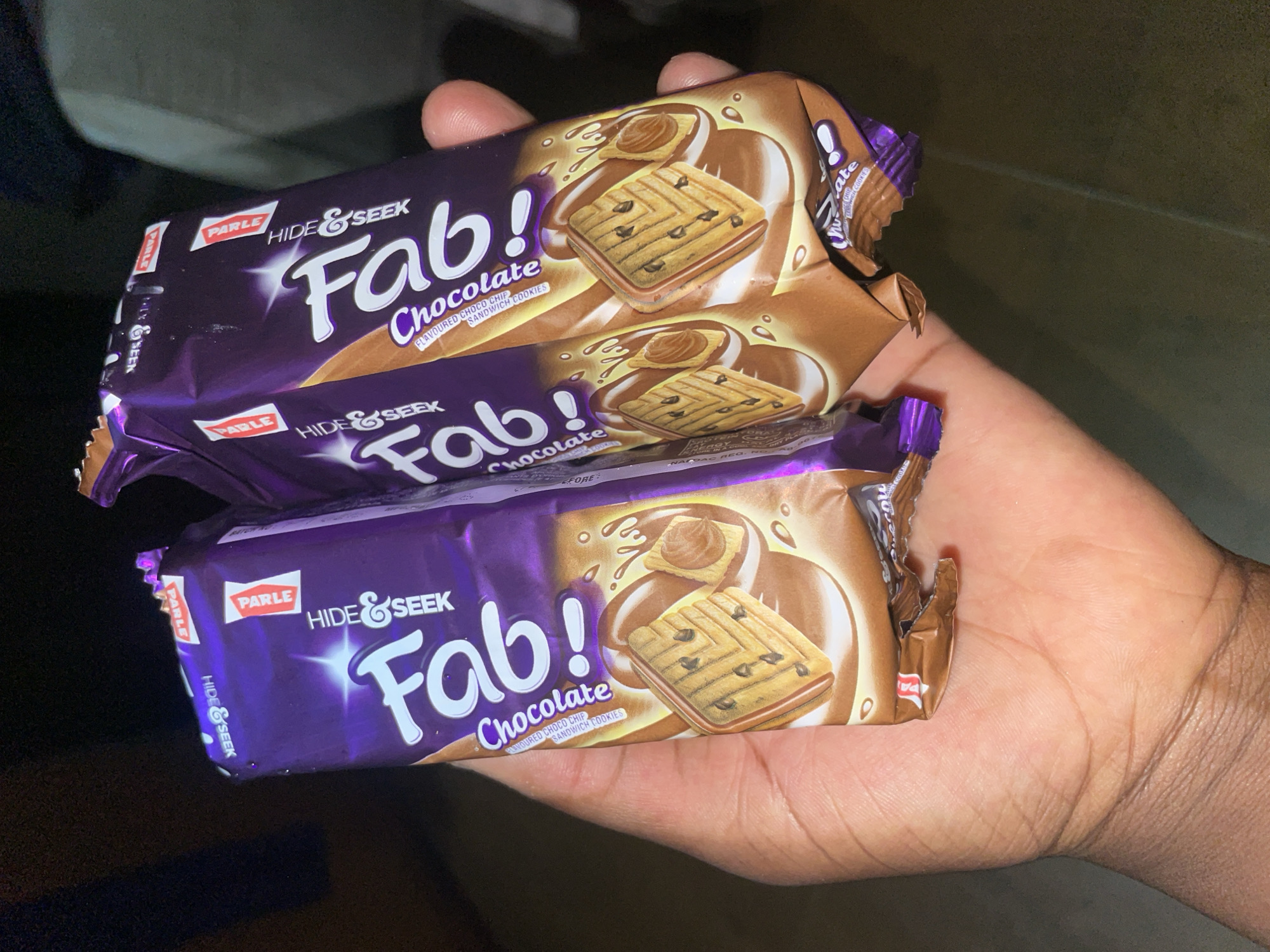 A human hand holds three unopened packages of "Parle Hide & Seek Fab! Chocolate Flavoured Choco Chip Sandwich Cookies." The location is unknown and cannot be determined from the image. The city of Kurunduma, Nigeria cannot be verified as the location from the image.