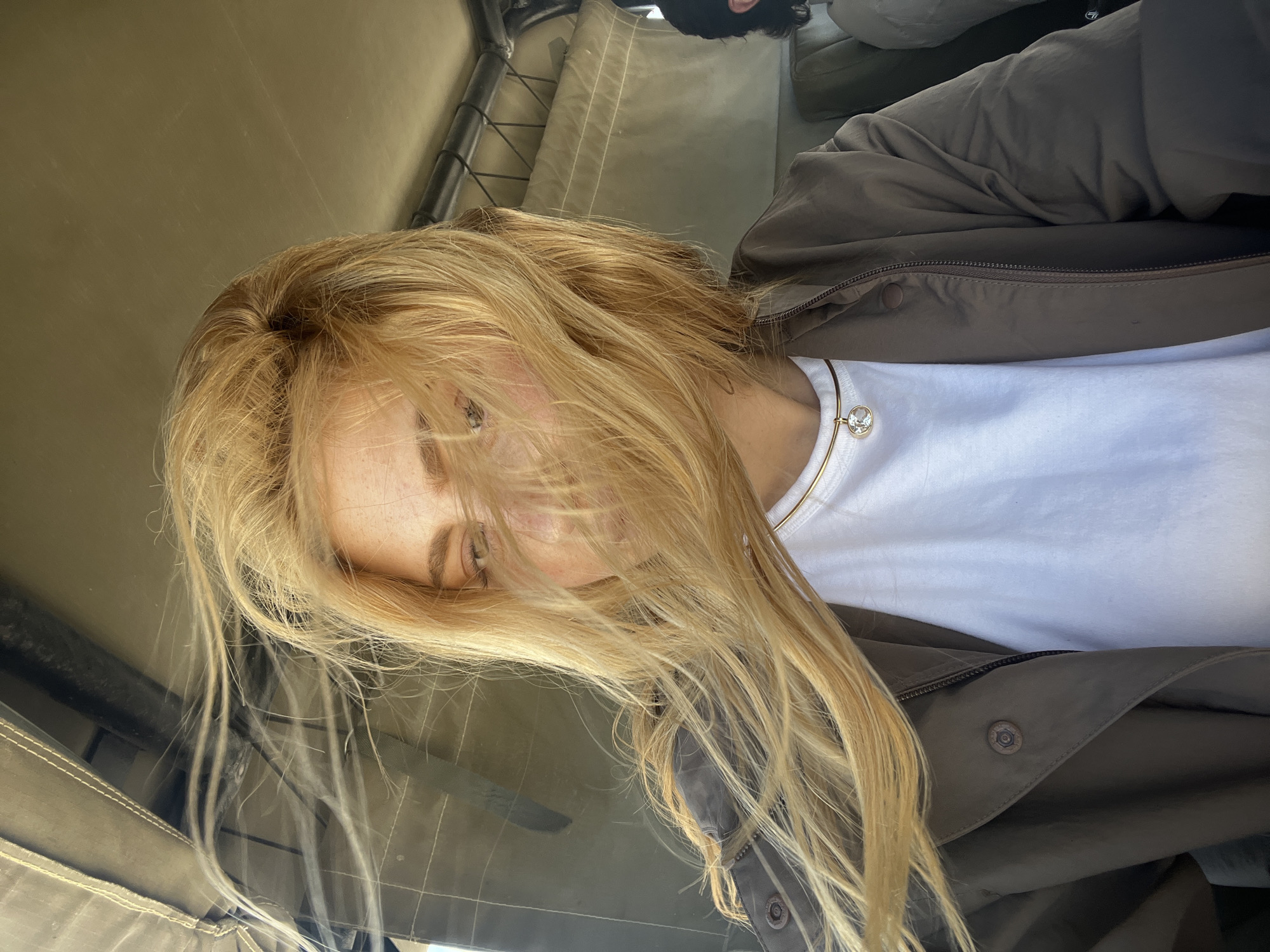 A woman with long blonde hair, wearing a white t-shirt and olive-green jacket, is inside a vehicle. The interior of the vehicle appears to be canvas or covered in a similar material. A gold necklace with a crystal charm is visible. The location is implied to be Gucta, Botswana, based on the prompt. The scene suggests the subject is in a safari vehicle or similar transport.