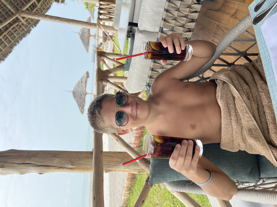 A young male is seated on a shaded outdoor patio overlooking a beach and ocean on the island of Zanzibar, Tanzania. The individual, appearing to be an adolescent, has short, light brown hair and wears aviator-style sunglasses with dark lenses. He is shirtless, with a light brown towel wrapped around his lower body. He holds a tall glass in each hand, both containing a dark beverage, ice, a lemon slice, and a red straw, extending them forward with a smile directed towards the camera. He occupies a grey-colored chair with a woven back and a teal cushion. A wooden table with a light blue placemat and silverware is partially visible in the foreground. The patio structure features rustic wooden posts and railings, supporting a thatched roof. Beyond the railing, a strip of green vegetation borders a sandy beach. The ocean, characterized by calm, light blue water, extends under a light blue sky. Several thatched beach umbrellas are present on the sand in the mid-ground.