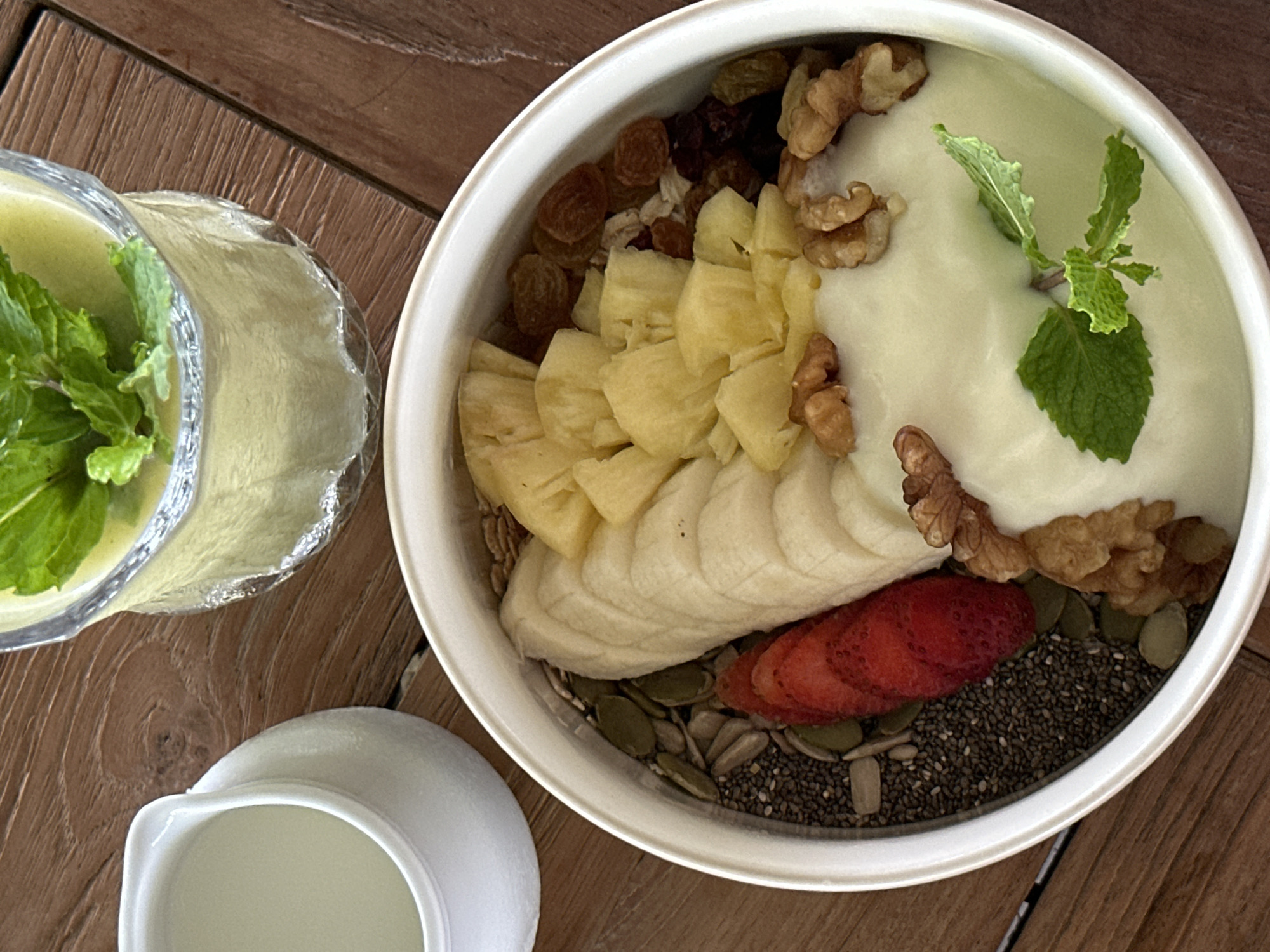 A top-down view showcases a white ceramic bowl filled with a breakfast bowl creation set on a wooden surface. The bowl contains a base of chia seeds, pumpkin seeds, and granola, upon which are layered slices of banana and strawberry, chunks of pineapple, raisins, and walnuts. The bowl is topped with a creamy white yogurt, garnished with a sprig of fresh mint. Beside the bowl rests a glass pitcher containing a green liquid, also garnished with mint, and a small white ceramic pitcher filled with a light-colored liquid, potentially milk or a smoothie base. The scene is likely photographed indoors, possibly in a cafe or personal dining area, in Pekutatan, Indonesia.