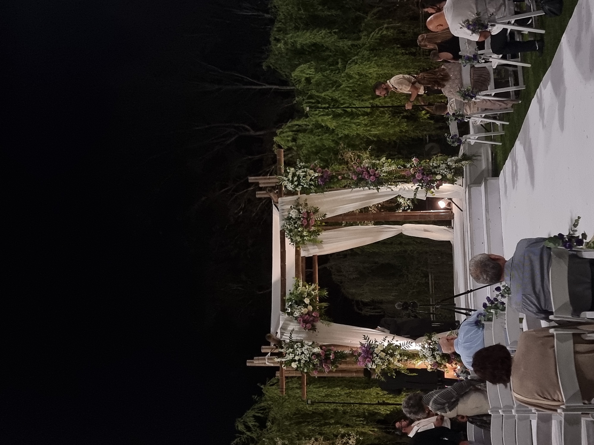 This image depicts an outdoor evening wedding ceremony in Ga'ash, Israel. The scene is illuminated by artificial lighting, suggesting it is nighttime. The setup includes a decorative archway adorned with white fabric and floral arrangements in shades of purple and white. Rows of white chairs are set up on either side of a white aisle, indicating seating for guests. Several people are visible, some seated and others standing, appearing to be attendees or participants in the ceremony. One individual, a man with a light-colored shirt, is seen standing and facing the archway, possibly officiating or involved in the proceedings. The presence of a microphone stand near the archway further suggests a formal event like a wedding. The background features lush greenery and trees, characteristic of an outdoor venue. The overall atmosphere appears to be solemn and celebratory, typical of a wedding event. No visible text is present in the image.