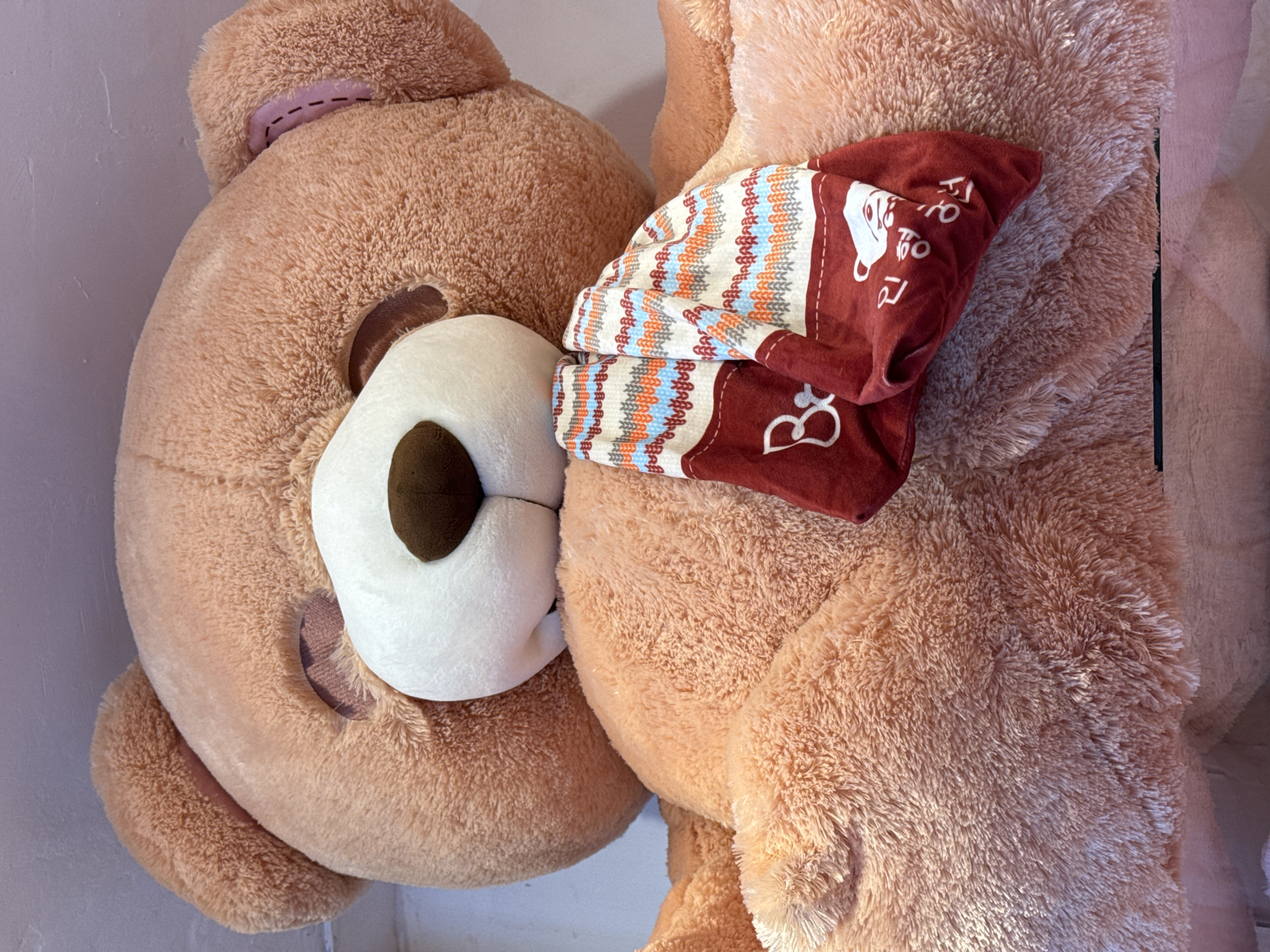 The image shows a close-up view of a large, plush teddy bear. The teddy bear is a light brown color and appears to be very soft. It is wearing a red scarf with a patterned design. The scarf has white, orange, blue, and gray zigzags on a white background. There are also some Korean characters on the red part of the scarf. The teddy bear's face is visible, with a white muzzle, a brown nose, and closed eyes. The bear is positioned in a way that suggests it is sitting or lying down, with its arms resting in front of it. The background is a light-colored wall. The image is taken from a low angle, looking up at the teddy bear. There is no specific indication of a location in Denpasar, Indonesia within the image itself.