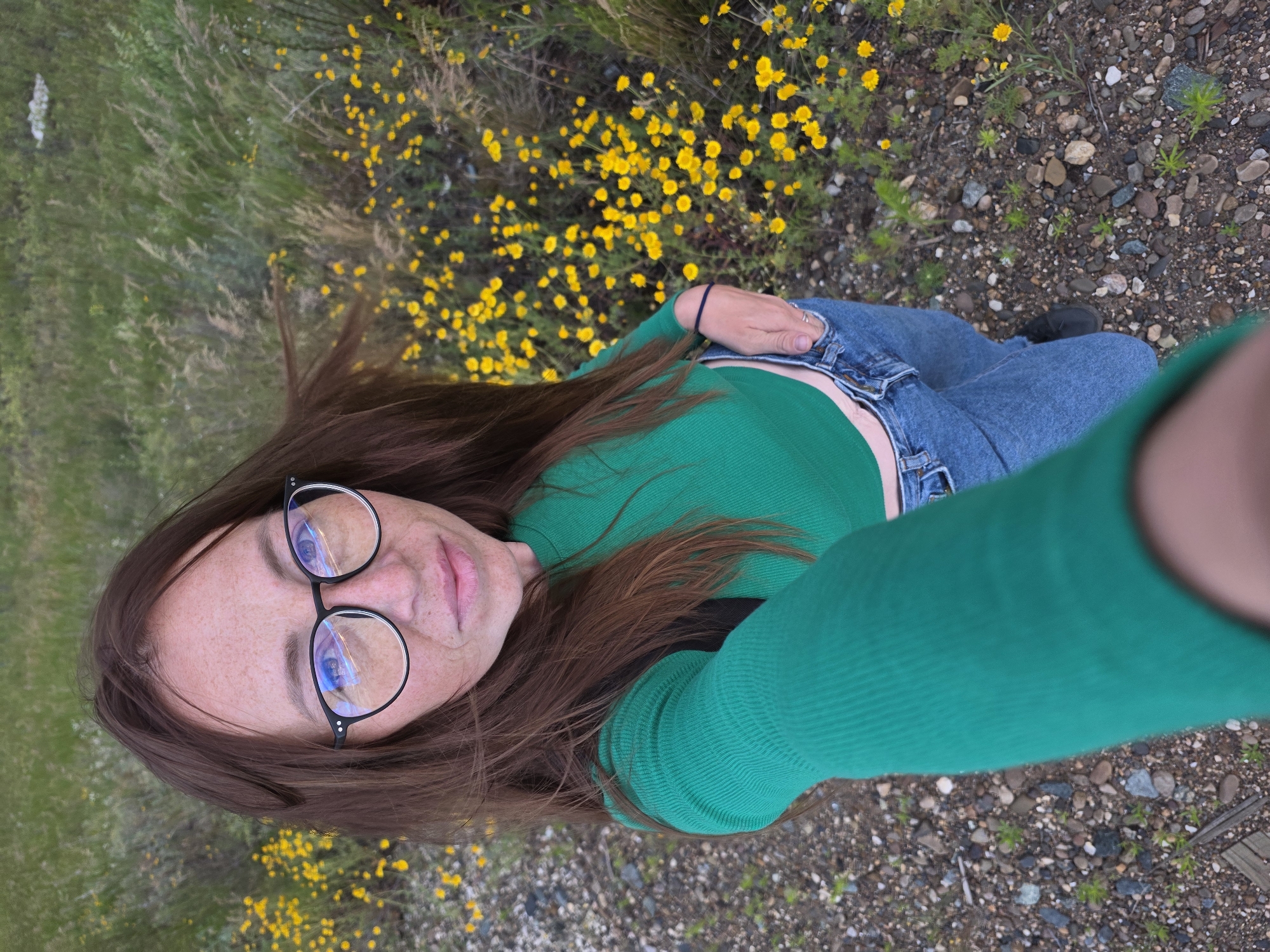 A person with long brown hair and freckles is taking a selfie outdoors. They are wearing glasses with black frames, a bright green long-sleeved shirt, and blue jeans. The person is sitting on the ground amidst a field of small yellow wildflowers and gravelly dirt. The background shows more green grass and some scattered rocks. The lighting suggests it is daytime, possibly overcast. The person's expression is neutral, and they appear relaxed. There is no visible text in the image. The image appears to be taken in a natural, outdoor setting, perhaps a meadow or a roadside with wildflowers.