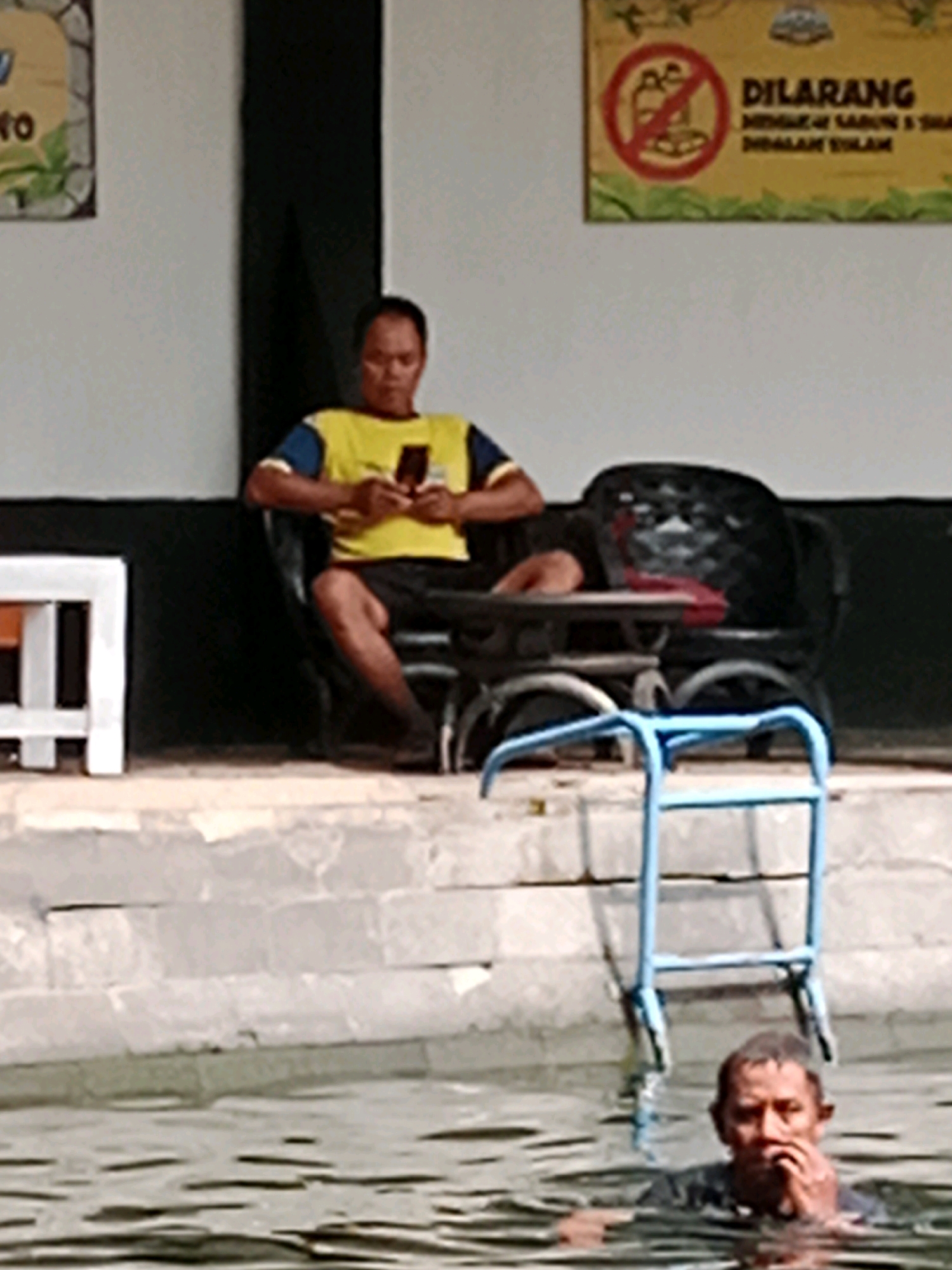 In Jentekan, Indonesia, a man sits in a black chair with a phone in his hand, wearing a yellow shirt and black shorts. Next to him is a white table with a dark object on it. In the background, a sign is visible with the word "DILARANG" and an image of a crossed-out soap. In the foreground, a man is partially submerged in water, near a blue ladder.