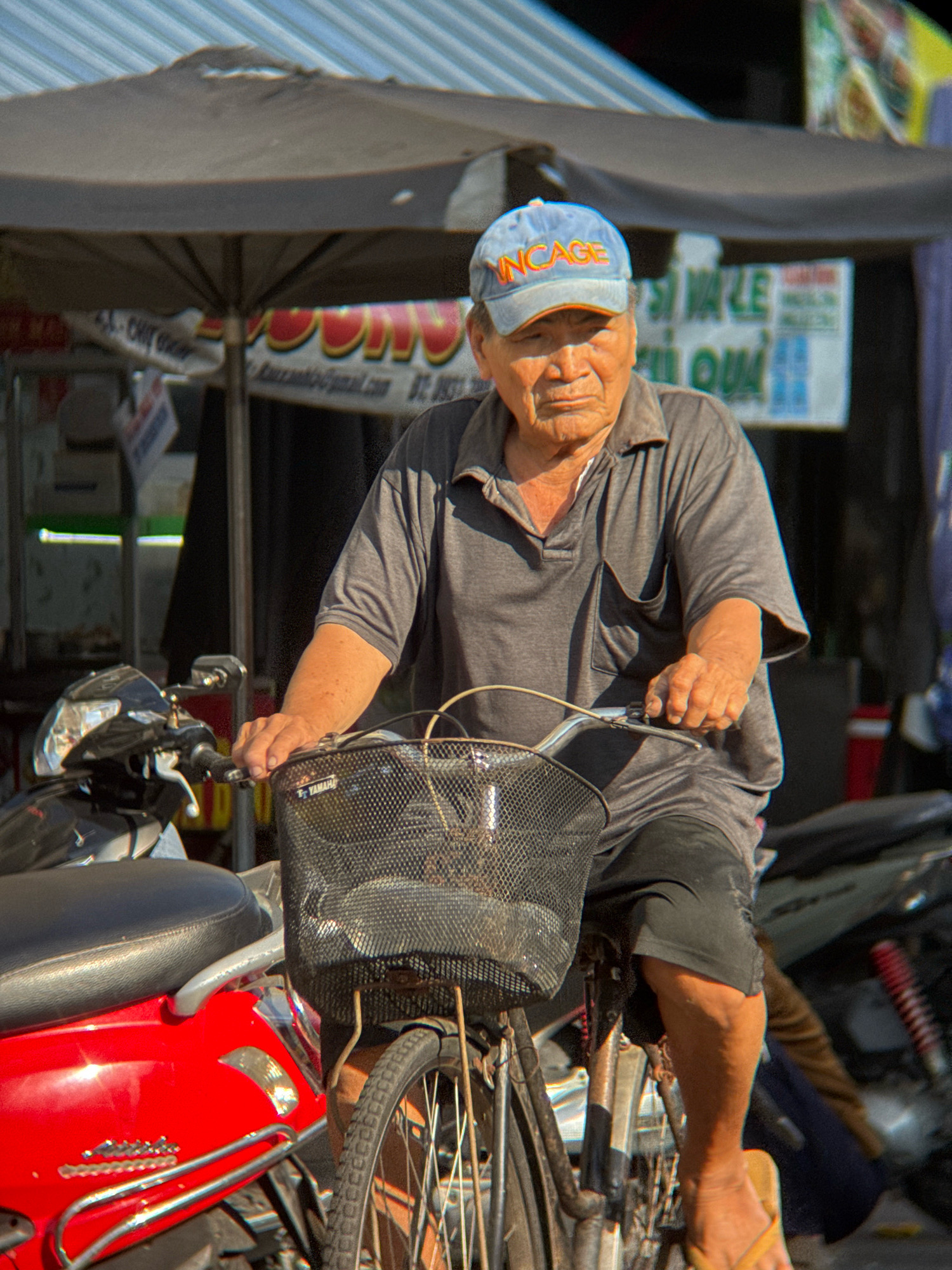 In Nha Trang, Vietnam, an elderly man wearing a light blue cap with orange lettering that reads "INCAGE" and a grey polo shirt is riding a bicycle. The man has wrinkled skin and is looking downwards, with his face partially obscured by shadow. He is holding the handlebars of the bicycle, which has a black mesh basket attached to the front. The bicycle is red and black. In the background, a striped awning casts a shadow over the scene, and signs with Vietnamese text are visible. The environment appears to be a street or market.