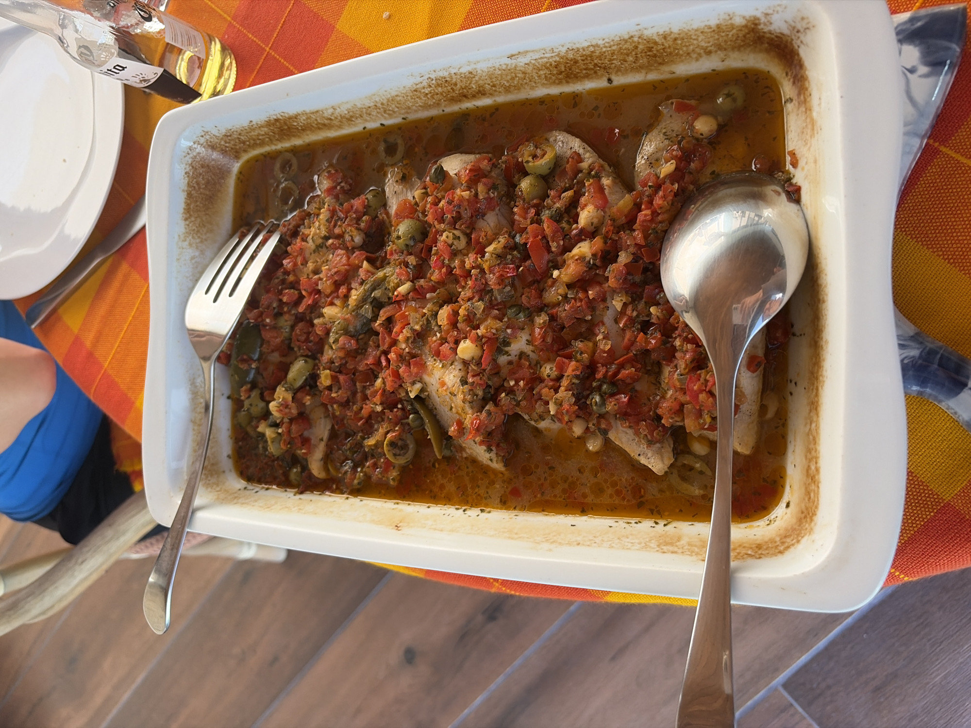 A rectangular white baking dish filled with cooked fish, possibly cod, covered in a red tomato-based salsa with green olives and capers is the focus of the image. The dish sits on an orange and yellow checkered tablecloth, with a silver fork and spoon positioned on either side. A white plate and bottle are also visible to the left, partially obscured. The scene is captured indoors, likely a dining area. The image was captured at an elevated angle. I have no information on the location in San Bernardino Tlaxcalancingo, Mexico.