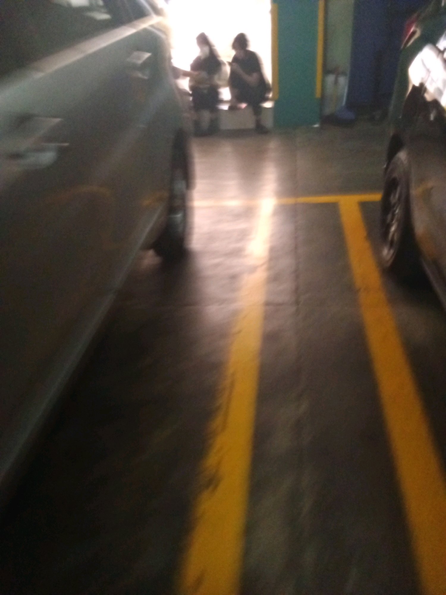 This is a slightly blurry, low-angle shot taken in what appears to be an indoor parking garage or lot with a concrete floor marked by yellow lines. In the foreground on the left is the side of a silver car. In the middle ground, two individuals are seated on a low bench. The person on the left is partially obscured by the car but appears to be wearing dark clothing and a light-colored scarf or head covering. The person on the right is also wearing dark clothing and is seated with their legs drawn up, looking at a handheld device. Behind them, there is a wall that is painted green and blue. The lighting suggests an indoor space with artificial lights. There are no clear indicators of the specific location within Bandung City, Indonesia, but the setting suggests a public or commercial area like a mall, office building, or residential complex parking facility.