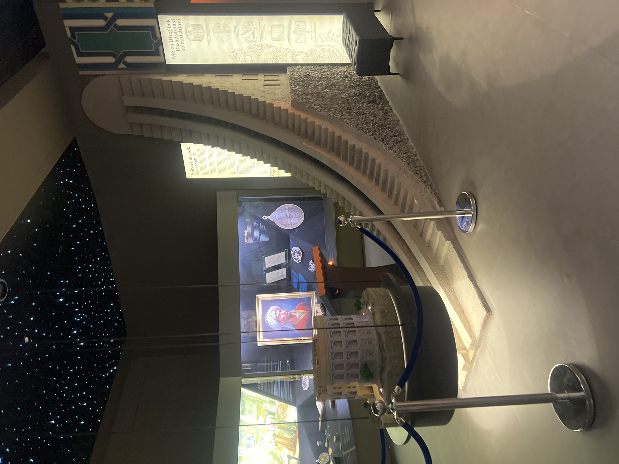 The image depicts an indoor exhibition hall, likely a museum, featuring models and informational displays. The ceiling is dark and adorned with numerous small, bright lights resembling stars. In the foreground, a velvet rope stanchion stands on a metallic base, with a blue rope extending to the left. To the left of the stanchion is a circular display containing a miniature model of a circular, multi-story building with arched windows, situated on a textured base.
Dominating the mid-ground and right side of the image is a large, detailed model of a curved structure with numerous steps ascending it, appearing to be an observatory or astronomical instrument. The lower portion of this model has a rough, stone-like texture. Adjacent to this model, to the right, is a dark upholstered bench with metal legs.
Behind the models, display cases are visible. One case contains a framed portrait of a man wearing a turban and a red garment. Other displays show what appear to be artifacts, scientific instruments, and printed information panels. One panel, in particular, has text in a foreign language and is illuminated. The overall lighting is dim, with spotlights illuminating the exhibits. The walls are dark grey, with some sections featuring decorative geometric patterns. The floor is a smooth, polished grey surface.