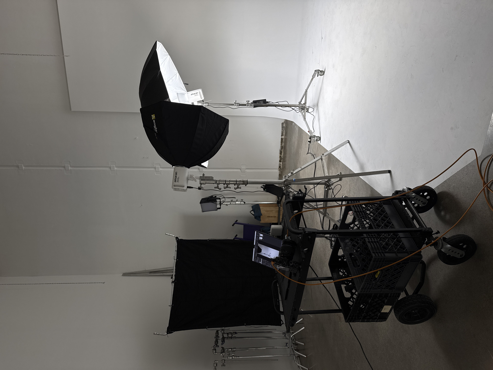The image shows a studio setup in what appears to be Los Angeles, United States of America. Dominating the scene is a large umbrella-style light modifier with a black exterior and a white interior, mounted on a stand. Adjacent to it is a black backdrop or flag, also mounted on a stand. A cart filled with equipment including a camera and monitor sits in the foreground. The environment is a white studio, likely a sound stage or photography studio. The scene suggests a film or photography shoot in progress or set up and waiting for use.