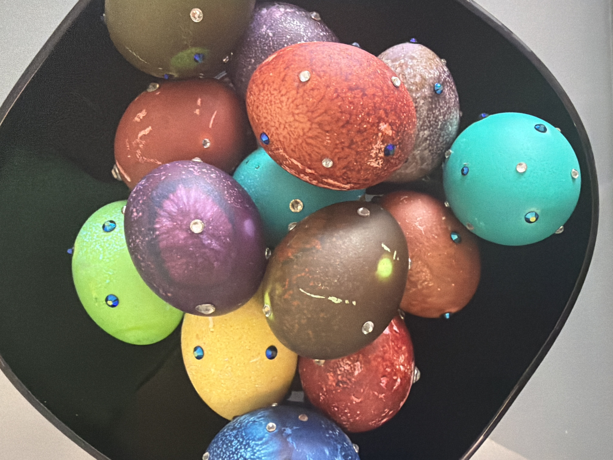 A close-up, high-angle shot depicts a black bowl filled with a collection of decorated Easter eggs. The eggs vary in color, including shades of green, yellow, brown, red, purple, blue, and turquoise. Each egg is adorned with small, shiny embellishments, possibly rhinestones or other decorative stones, placed on its surface. The bowl is situated against a background that is light grey or white, which is out of focus. The exact location is not ascertainable.