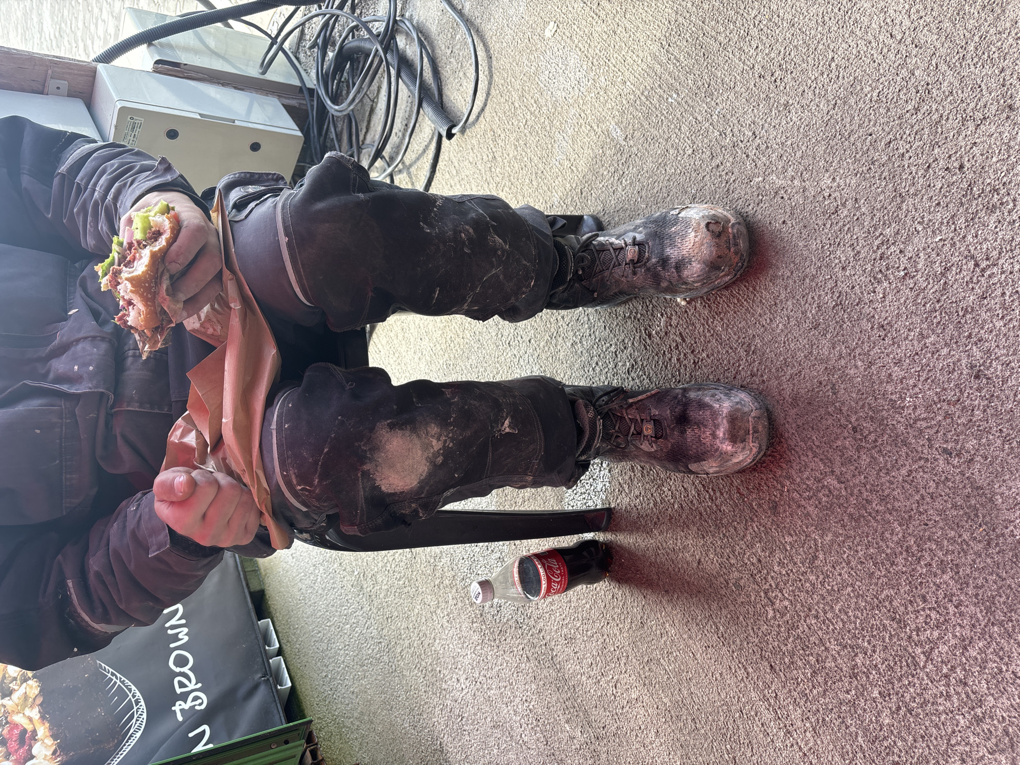 A person wearing dirty work clothes and work boots is eating a sandwich while standing outside. A partially empty bottle of Coca-Cola lies on the ground next to the person's feet. Part of a sign is visible in the lower left corner, including the words "N BROWN." The location appears to be an outdoor urban setting, possibly in Burgas, Bulgaria. Wires and electrical boxes are visible in the background.