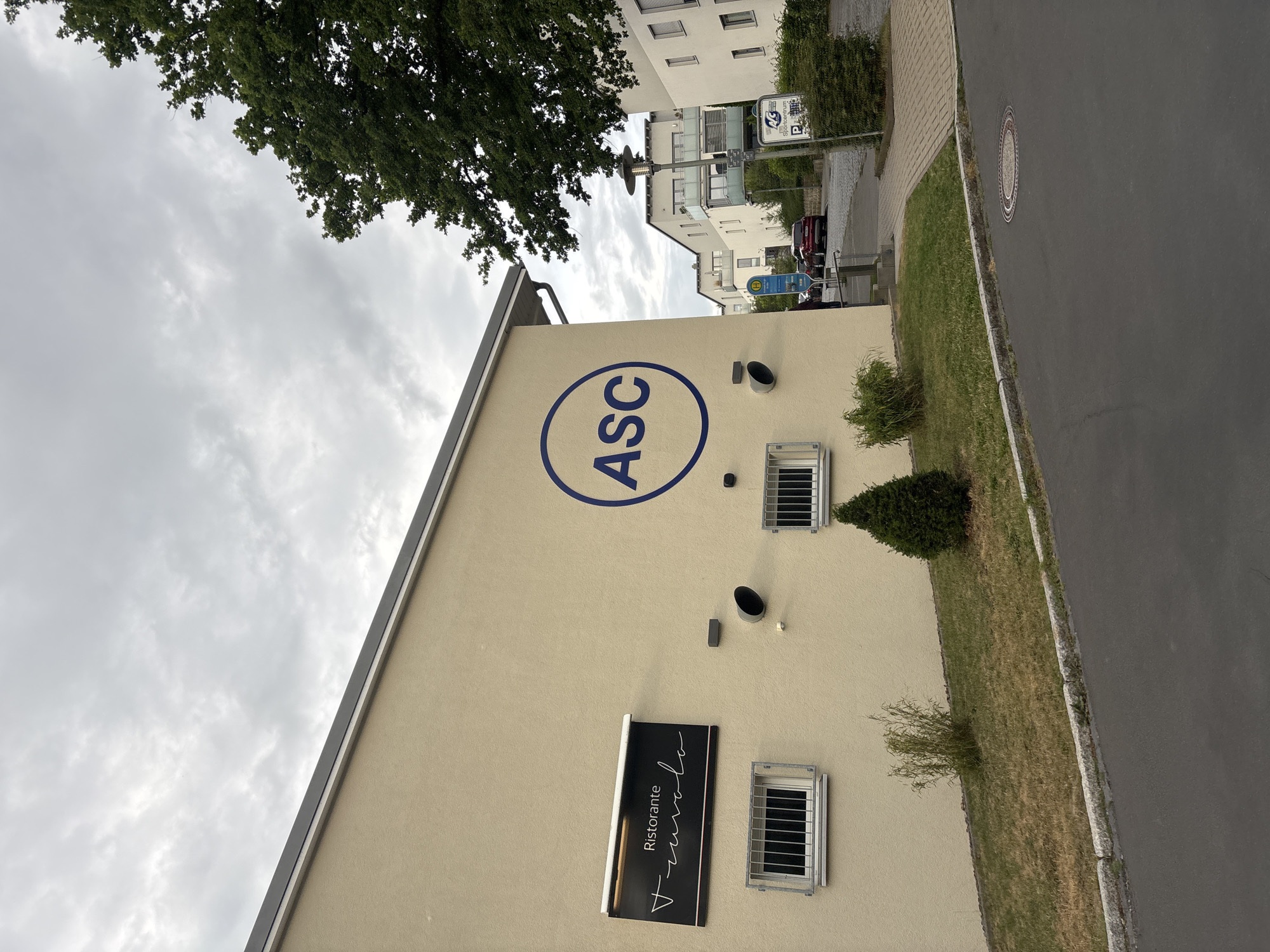 The image shows the exterior of a building with a beige stucco finish. A sign for "Ristorante TraVolo" is mounted on the left side of the building. To the right of this sign, a large blue circle with "ASC" in white letters is painted on the wall. There are two barred windows on the lower portion of the building, and a small conical evergreen tree is planted in front of the right window. A pathway with cobblestones leads towards the right side of the frame, where there is a sign for "ASC Sportcentrum". The scene appears to be in a modern urban or suburban setting, with apartment buildings visible in the background. The sky is overcast. The image was taken in Göttingen, Germany.