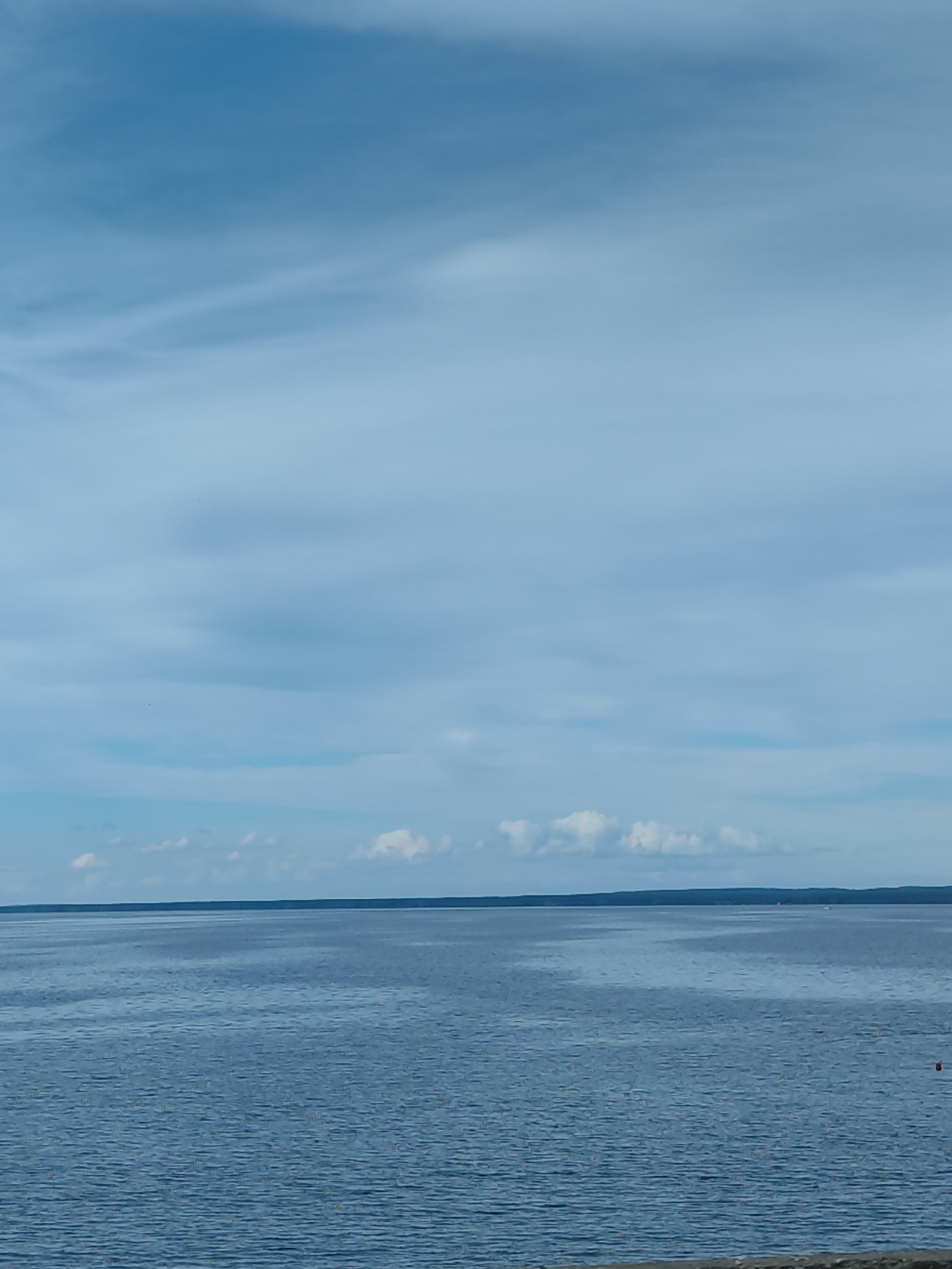 A wide shot captures a serene body of water under a vast, partly cloudy blue sky. The water is a deep blue, with subtle ripples creating texture across its surface. In the distance, a dark, flat horizon line suggests land or a distant shoreline. A few small white clouds are scattered in the sky, with some appearing wispy and stretched, while others are more defined. The overall impression is one of tranquility and open space, likely a view of a lake or the sea. A small red buoy is visible in the lower right corner of the image.