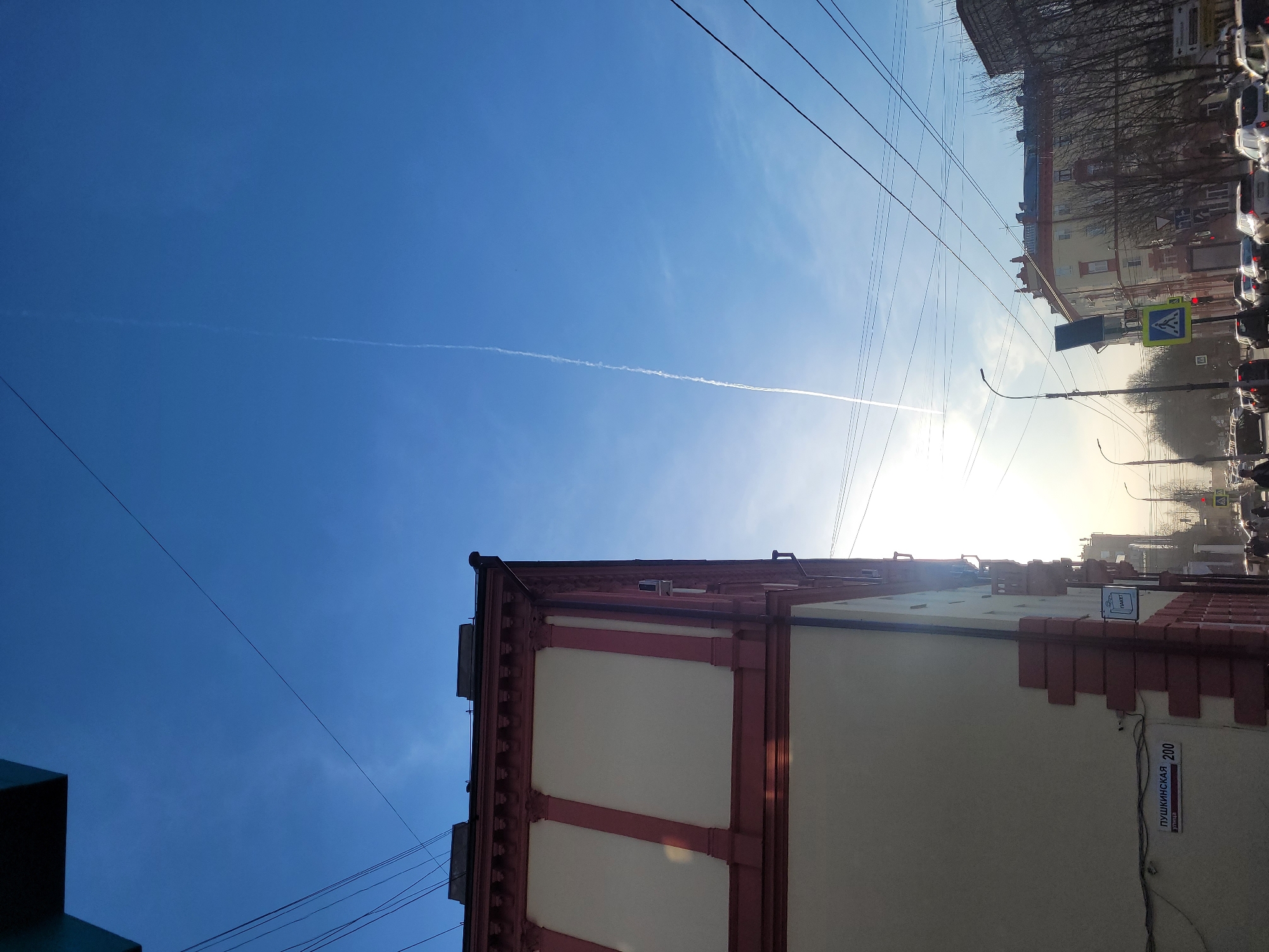 The image shows a low-angle, upward view of a city street in Izhevsk, Russia. The sky is predominantly blue with a contrail stretching across it. Overhead power lines crisscross the sky. A building with cream-colored walls and reddish-brown trim occupies the lower right portion of the frame. A street sign attached to the building reads "Пушкинская 200" indicating the location as 200 Pushkinskaya Street. Across the street, there are other buildings, trees without leaves, and parked cars. Pedestrian crosswalk markings and traffic signs are visible near a crosswalk. The sun appears to be low in the sky, casting a bright light through the scene. There are no visible people in the frame.