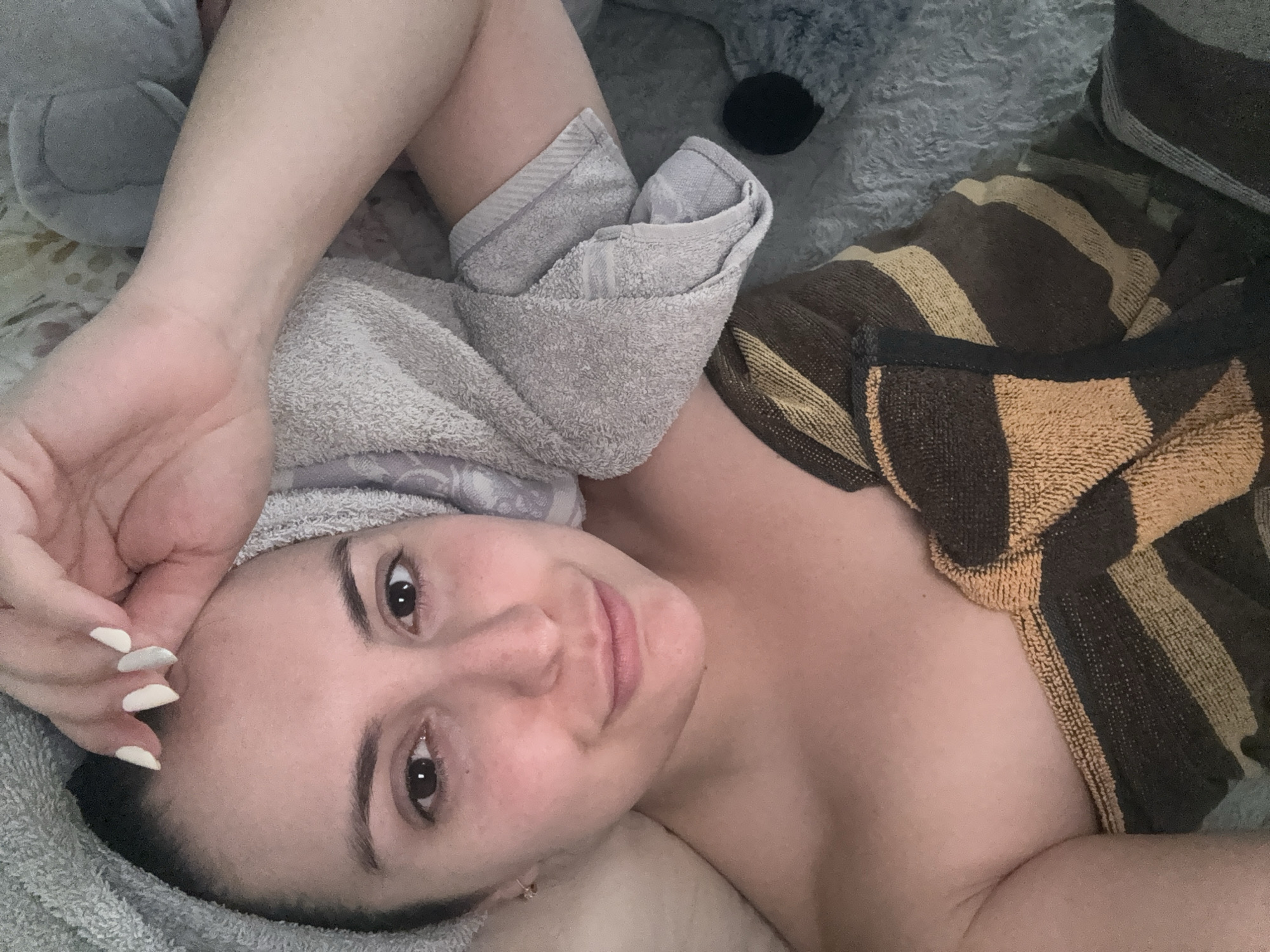 A woman is lying down with a gray towel wrapped around her head and a brown and tan striped towel covering her chest. She has a fair complexion, dark hair, and brown eyes. Her left hand rests on her forehead, and her nails are painted white. In the background, there are gray stuffed animals. There is no indication that the image was taken in Barcelona, Spain.