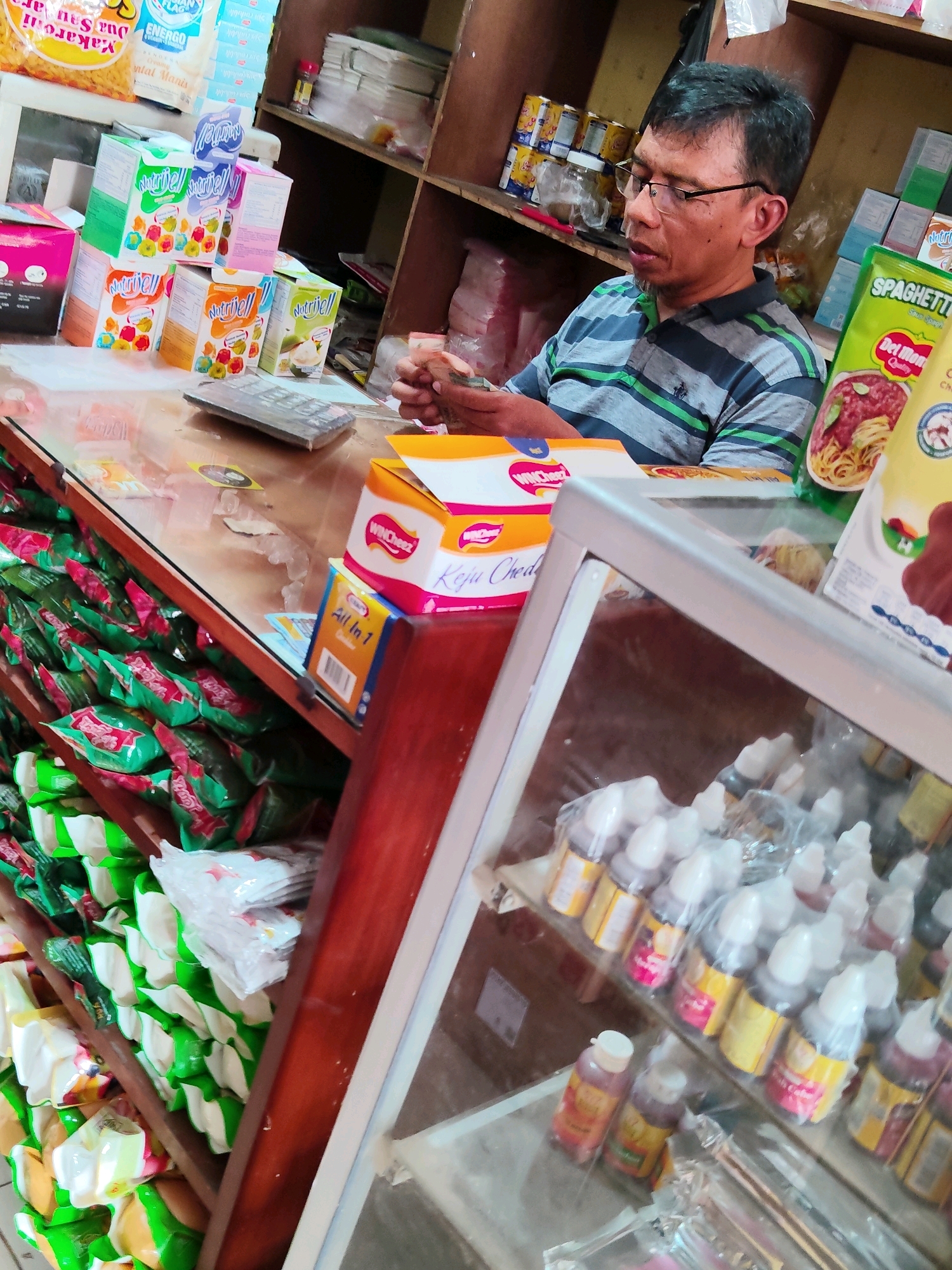 A man wearing glasses and a striped shirt is counting money at a counter in what appears to be a small grocery store or warung in Purwakarta, Indonesia. The counter is cluttered with various products, including boxes of instant pudding mix, packaged snacks, and small bottles of what may be food coloring or flavorings displayed in a glass case. Shelves in the background are stocked with canned goods, boxes of milk, and other packaged items. The overall lighting is dim, suggesting an indoor environment.