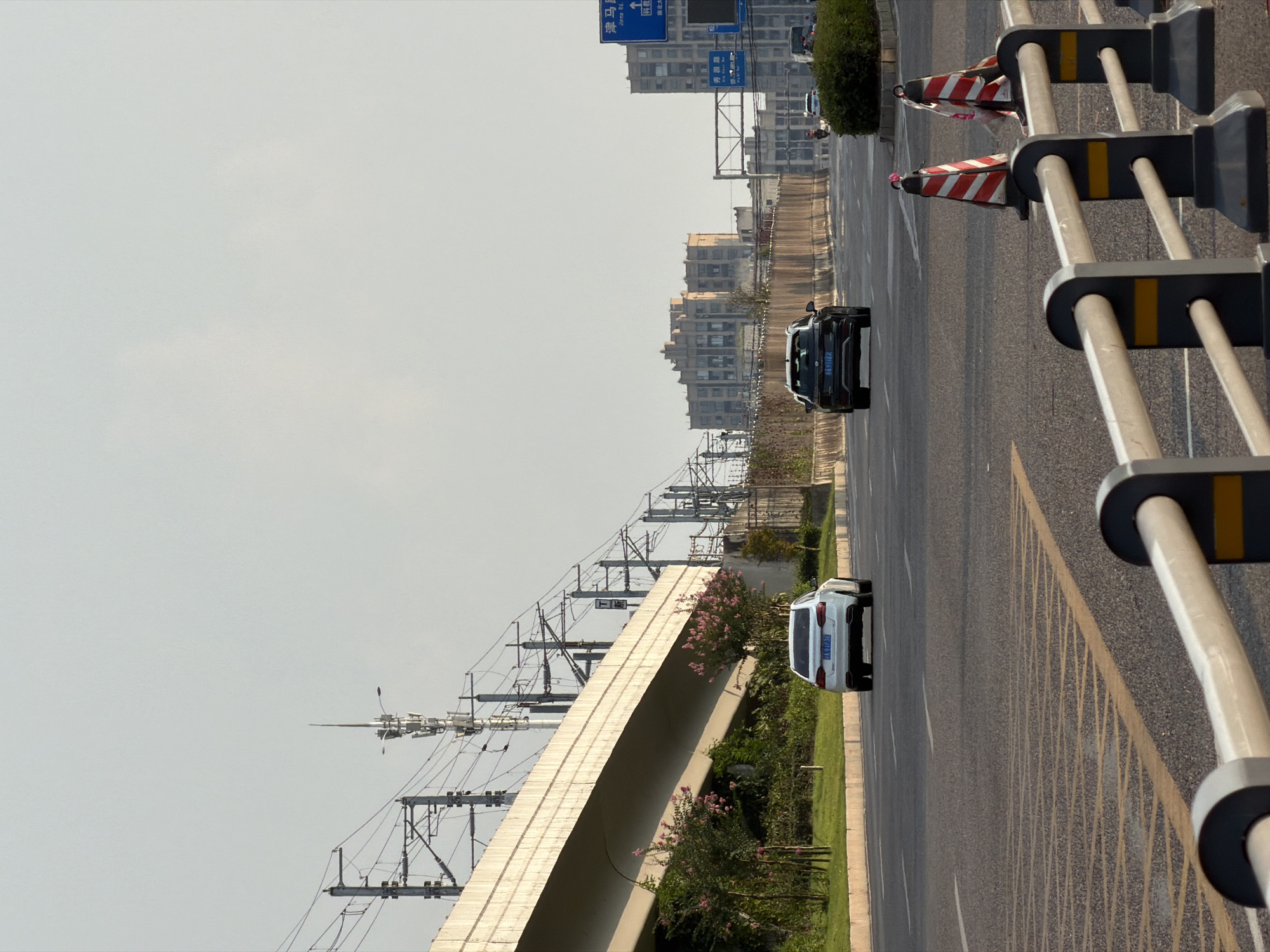 The image depicts a wide, multi-lane road in Jiangjin, China, under a clear, pale sky. A highway overpass with overhead railway lines runs from left to right across the upper portion of the frame. Several vehicles are visible on the road, including a white sedan and a black SUV. In the foreground, a barrier with reflective elements separates lanes, and traffic cones are placed on the roadside. The environment includes urban architecture in the distance, characterized by multi-story buildings. Greenery, including trees and bushes, lines the sides of the road. The overall scene suggests a typical urban traffic environment.
