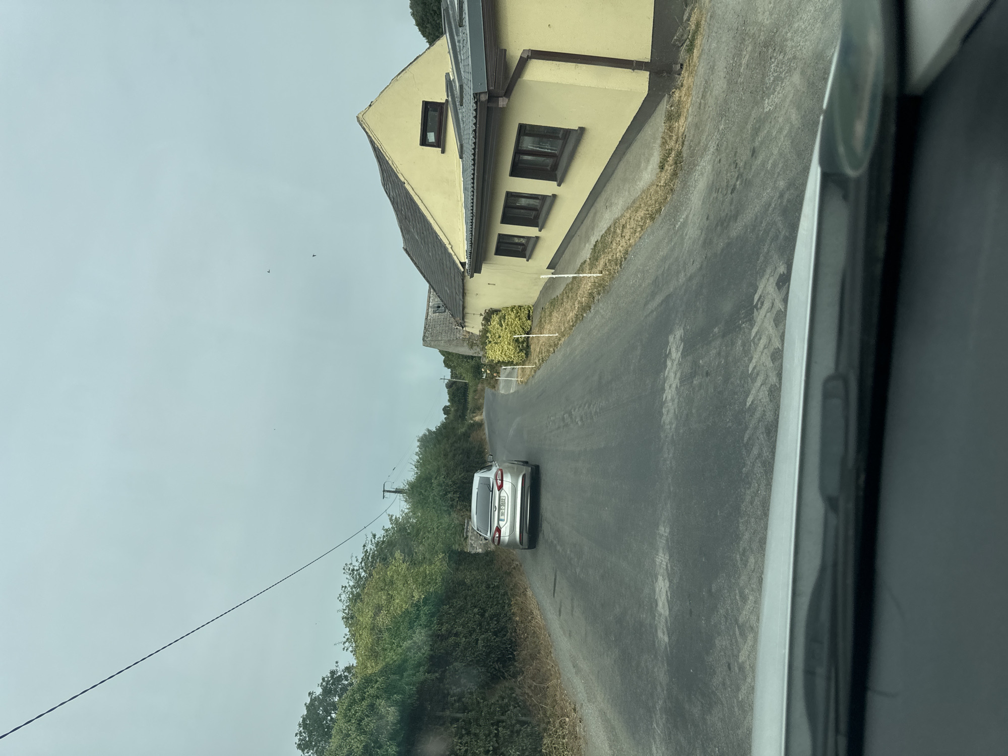A silver sedan is captured from a first-person perspective, driving on an asphalt road in what appears to be rural Ireland. The car is facing away from the camera, traveling uphill. To the right of the road is a long, single-story yellow building with a dark gray roof and brown trim. The building has several windows visible along its side. Further down the road, behind the yellow building, a portion of an older stone structure can be seen.
On the left side of the road, a dense green hedge lines the embankment, with trees visible beyond it. The sky is overcast and a light blue-gray. A thin power line stretches diagonally across the upper portion of the image. The foreground shows the edge of a car's dashboard and windshield wiper. The license plate of the silver sedan reads "191-LH-1022". The overall scene suggests a cloudy day in a semi-rural or suburban setting.