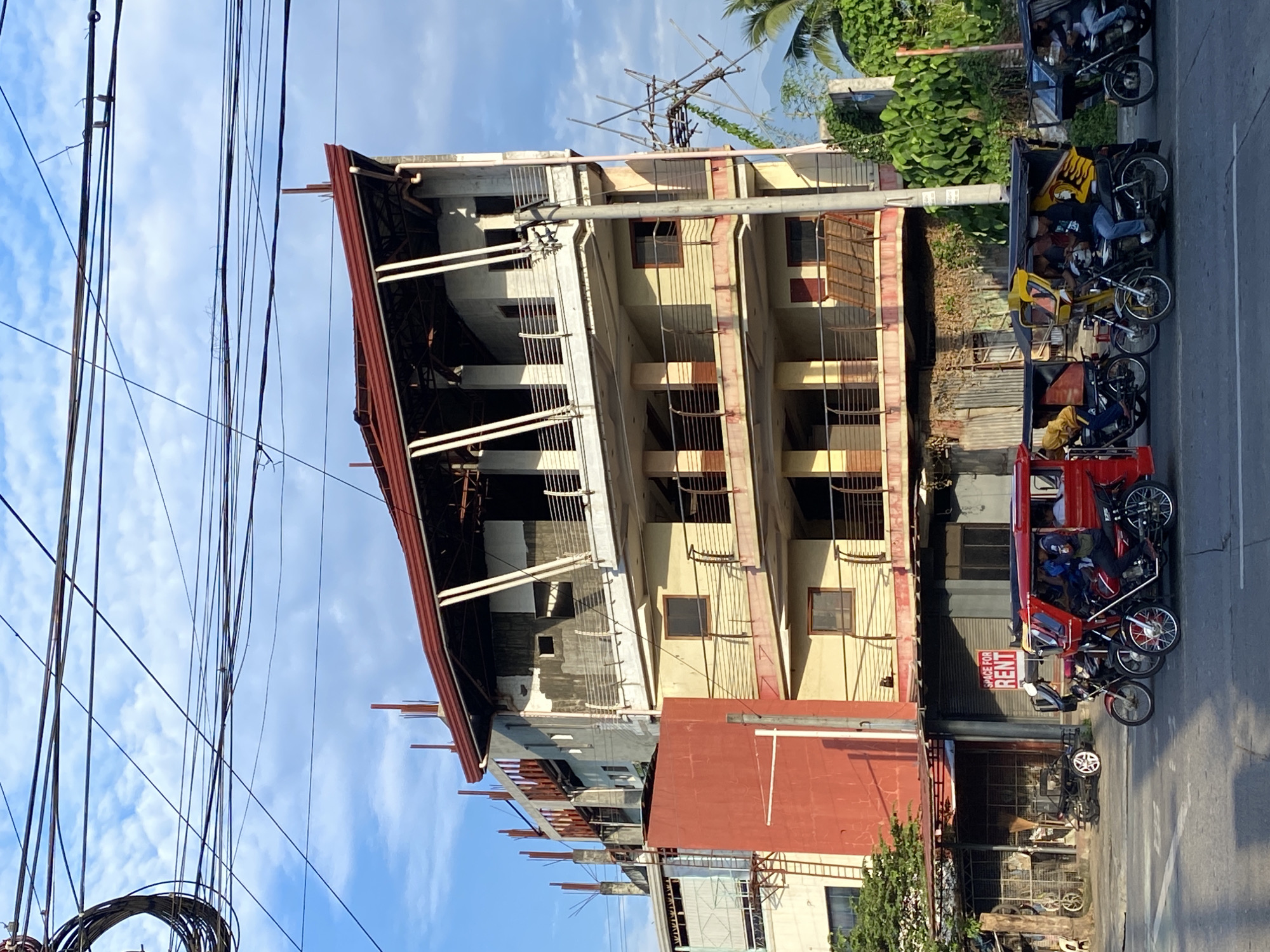 The image depicts a street scene in Davao City, Philippines, focusing on a multi-story building under construction. The building has a distinctive red-tiled roof and multiple levels with open balconies and unfinished facades. Below the construction site, a row of businesses can be seen, including one with a "SPACE FOR RENT" sign. Several vehicles are present on the street, including a red passenger tricycle in the foreground, with passengers inside. Other tricycles, some yellow and black, are also visible in traffic, some with drivers and passengers. Overhead, a complex network of electrical wires crisscrosses the sky. The lighting suggests it is daytime, possibly late afternoon, with the sun casting shadows on the street. Lush green vegetation is visible on the right side of the frame, with palm trees and other foliage indicating a tropical environment. The overall scene captures a slice of urban life in Davao City, characterized by ongoing development and local transportation.