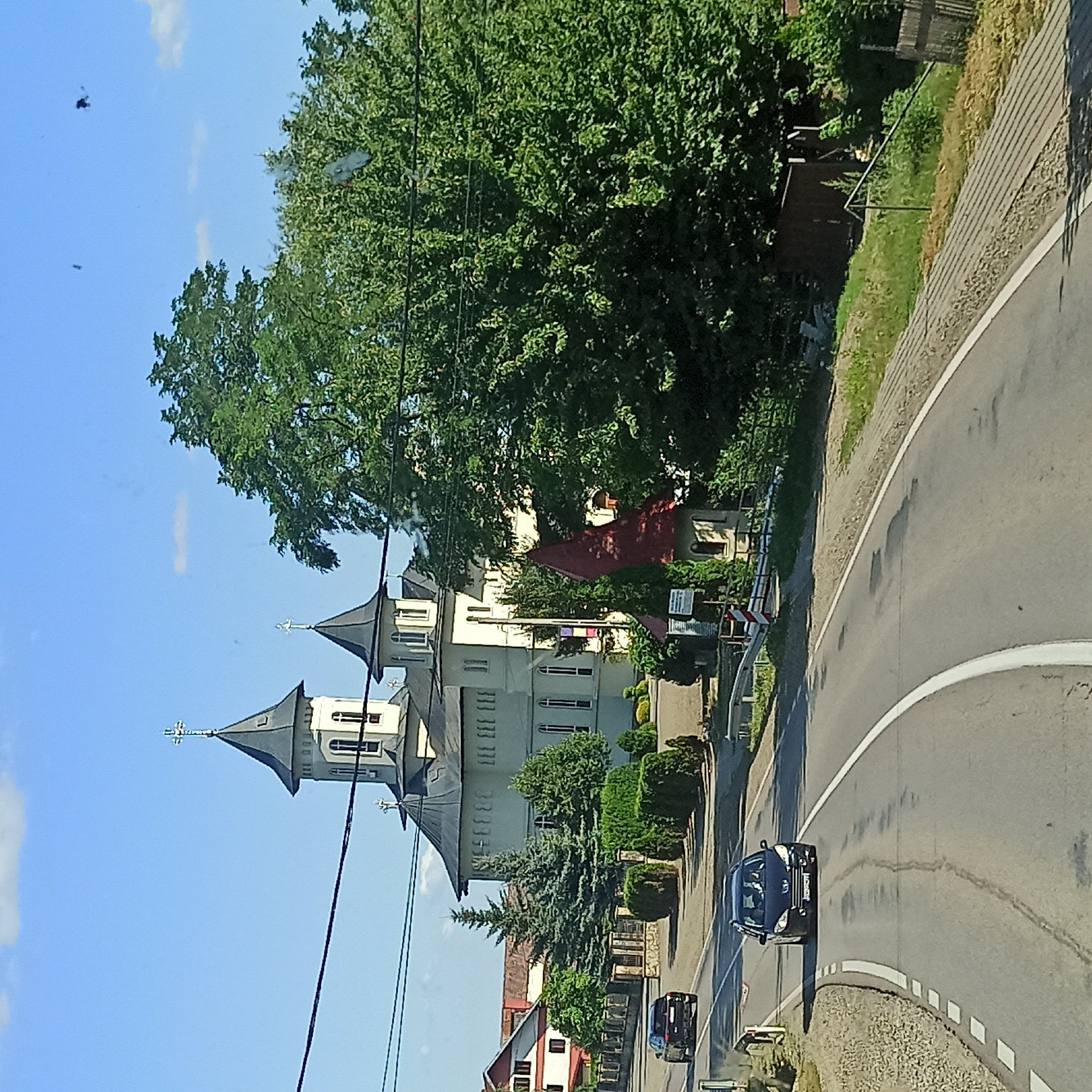 The image depicts a view of a street in Milișăuți, Romania, with a prominent church and lush greenery. A black Peugeot station wagon is driving on the road in the foreground, followed by another vehicle further back. The church, with its distinctive tall steeples topped with crosses, is situated to the left of a large, mature tree. Adjacent to the church, there are other structures and various types of trees and shrubs. The sky is blue with a few scattered clouds. A utility pole and wires are visible in the upper portion of the image. The road is paved and has white lane markings.