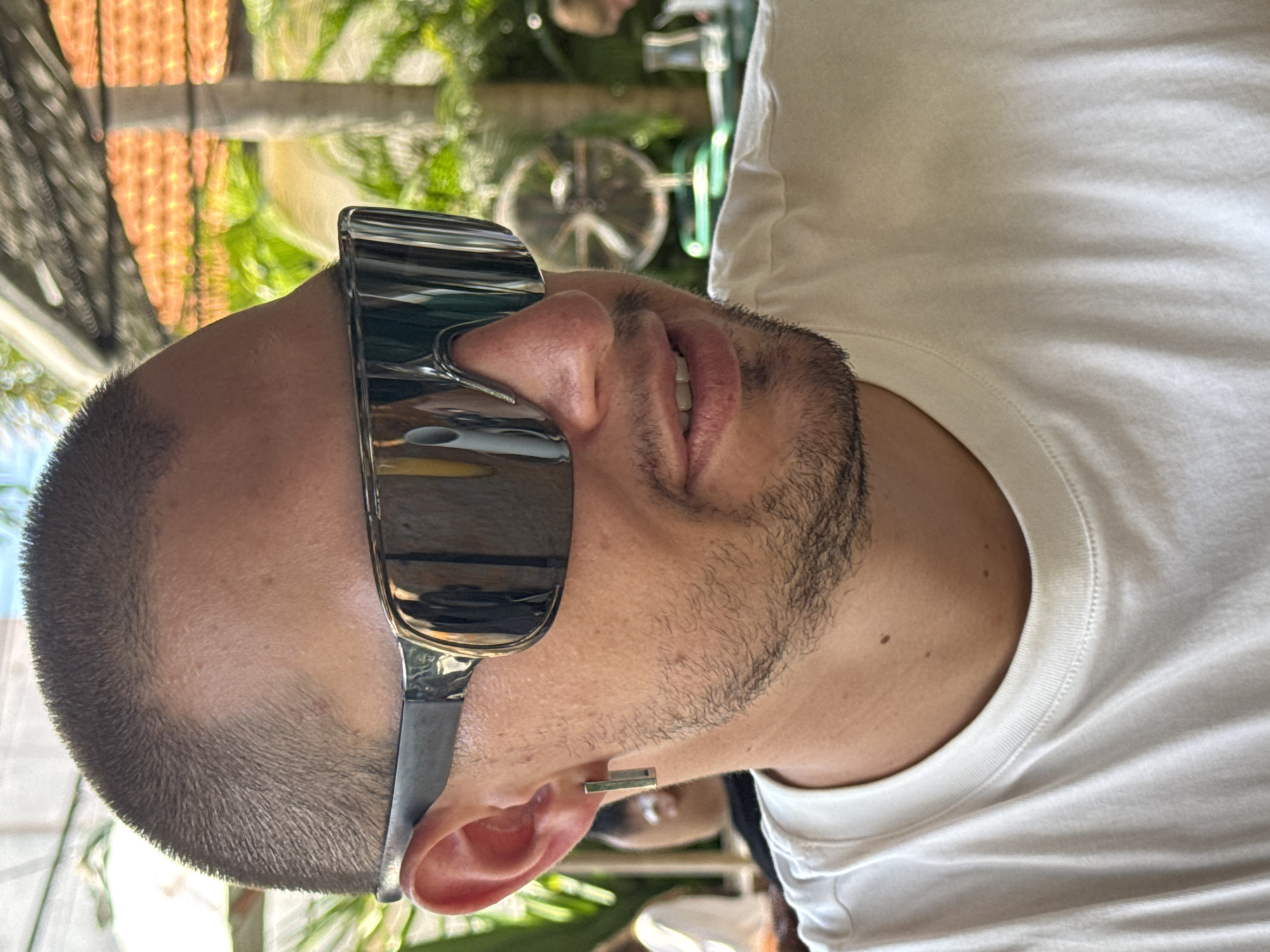 A man with a close-cropped haircut, beard, and sunglasses is wearing a white T-shirt. The setting appears to be outdoors, possibly in North Kuta, Indonesia, indicated by the foliage and building style in the background. There is a fan visible in the background, along with what appears to be an outdoor seating area. The man is facing partially towards the camera.