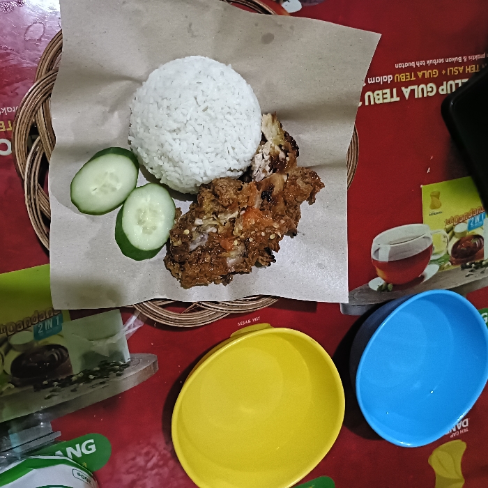 The image shows a plate of food on a red table in Tugu, Indonesia (latitude: -6.975, longitude: 110.332). The plate contains a serving of rice, two slices of cucumber, and a piece of fried chicken with a spicy sauce. The plate is on top of a woven bamboo placemat. There are also two empty bowls, one yellow and one blue, and a partial view of a glass with a brown liquid. The table is a vibrant red color, and the image appears to have been captured in a casual dining environment.