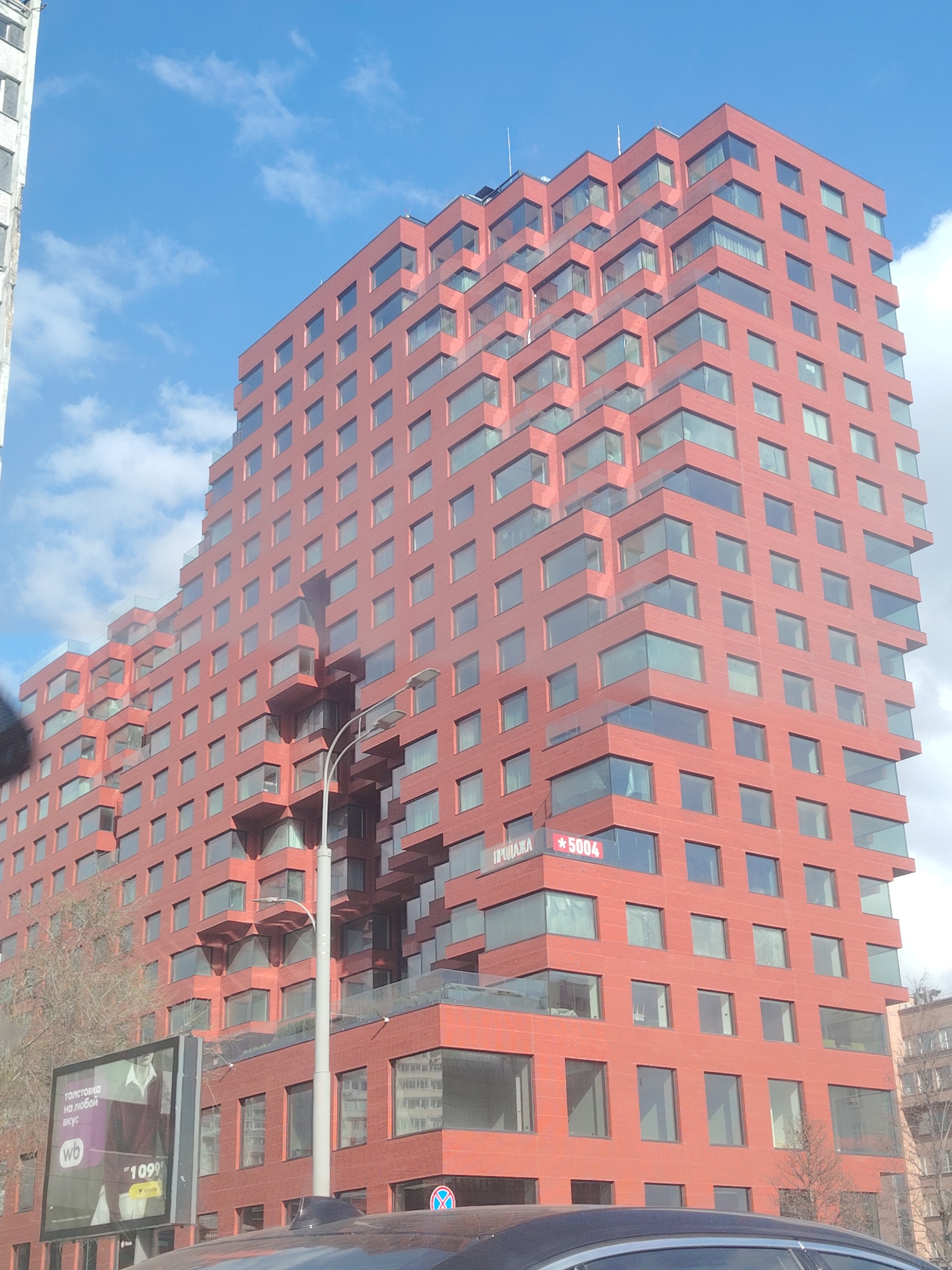 The image captures a modern, multi-story building with a distinctive stepped and terraced design, clad in a reddish-orange facade, likely brick or a similar material. Numerous windows of varying sizes dot its exterior, some featuring glass balconies, giving it a contemporary appearance. The building's architectural style creates a dynamic, receding profile towards the top.
A clear blue sky with scattered white clouds indicates a bright, sunny daytime scene. To the left, part of an older, lighter-colored building with visible wear is partially seen. A tall street lamp stands in the mid-ground, partially obscuring the red building. In the foreground, the dark roof of a car is visible, and on the lower right, a common Russian road sign (a red circle with a white bar, signifying "No Entry" or "No Stopping") is present.
Visible text on the red building, near one of the lower terraces, reads "ПРОДАЖА" (PRODAZHA, meaning "FOR SALE") followed by "*5004". Below, a large street billboard displays an advertisement for a "ТОЛСТОВКА НА ЛЮБОЙ ВКУС" (TOLSTOVKA NA LYUBOY VKUS, meaning "Hoodie for every taste"), featuring a person in a white shirt and a "wb" logo (likely Wildberries, a Russian online retailer) along with a price of "1099", presumably in Russian Rubles. The overall setting suggests a vibrant urban environment in Moscow, Russia.