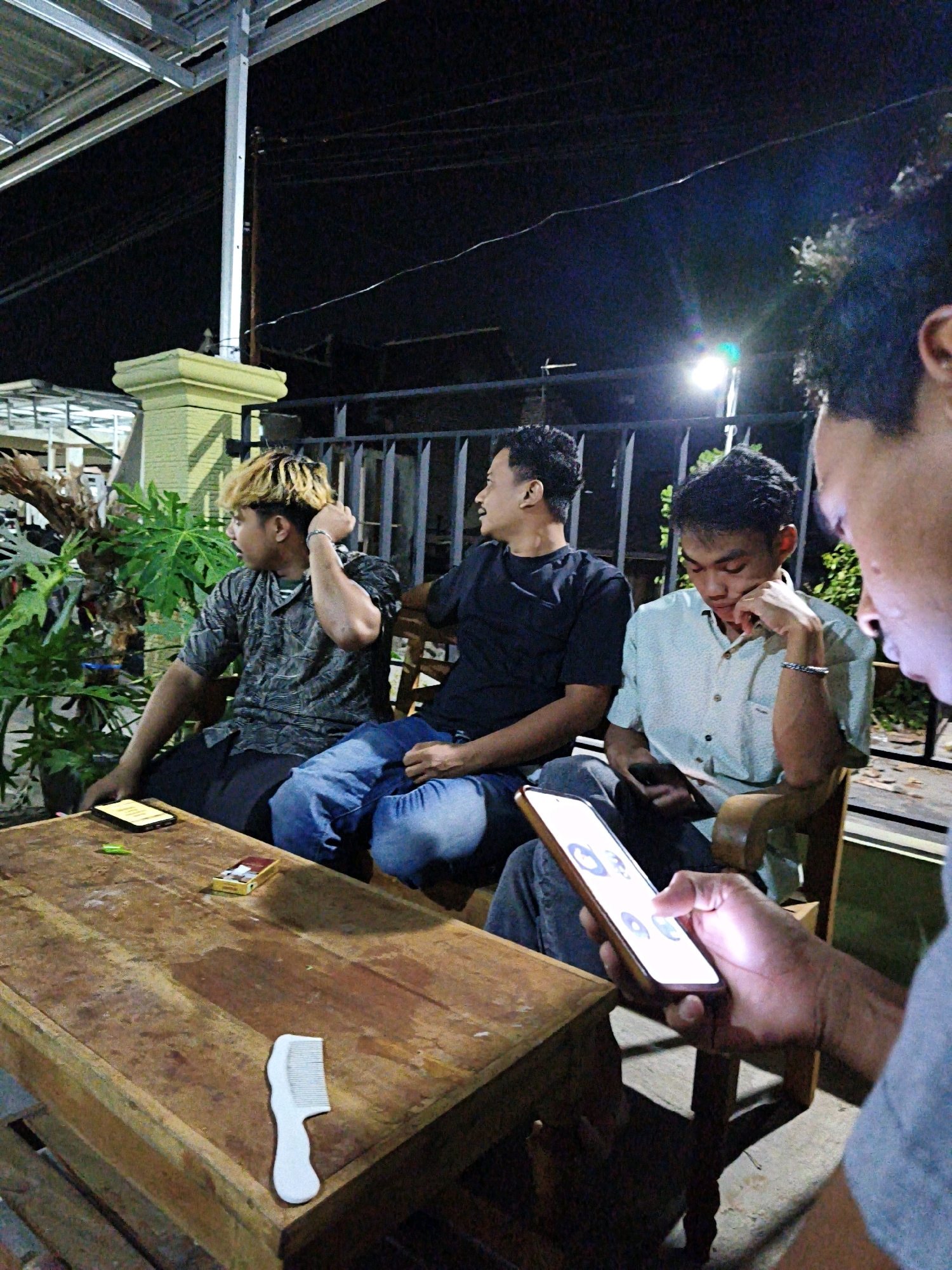 Four young men are gathered around a wooden table at night, likely at an outdoor cafe or gathering spot in Pengkok, Indonesia. The man in the foreground, facing away from the camera and to the right, is holding a glowing smartphone, his finger touching the screen. To his left, a man wearing a light blue collared shirt is also looking down at his phone, which is resting on his lap. Further left, a man in a black t-shirt is looking off to the side, away from the group. The man on the far left has blond and dark dyed hair, wears a patterned short-sleeved shirt, and appears to be adjusting something on his head. The table in front of them holds a pack of cigarettes, a lighter, and a white comb. The background features a dark night sky, some foliage, a fence, and structural elements of a building, including a metal roof and a pillar. The lighting suggests an artificial light source, possibly from the street or the cafe itself.