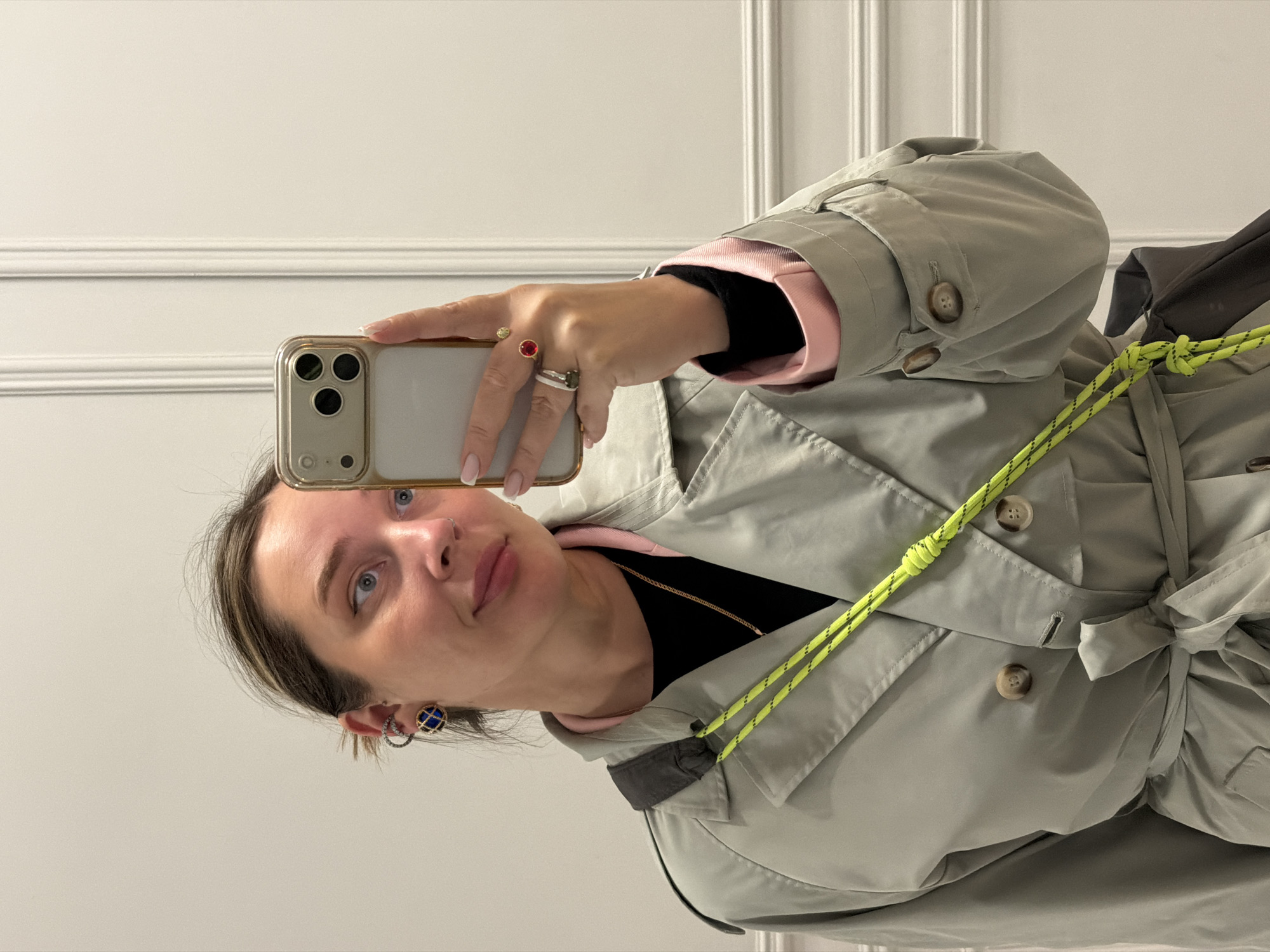 A woman is taking a selfie indoors against a white wall with decorative moldings. She is holding a smartphone in her right hand, visible near her face. She wears a beige trench coat with a neon yellow rope belt, over a black top with a pink collar and cuffs. The woman has short brown hair and blue eyes. She is wearing earrings and rings. The location is indoors, possibly in Moscow, Russia, based on the user's request.