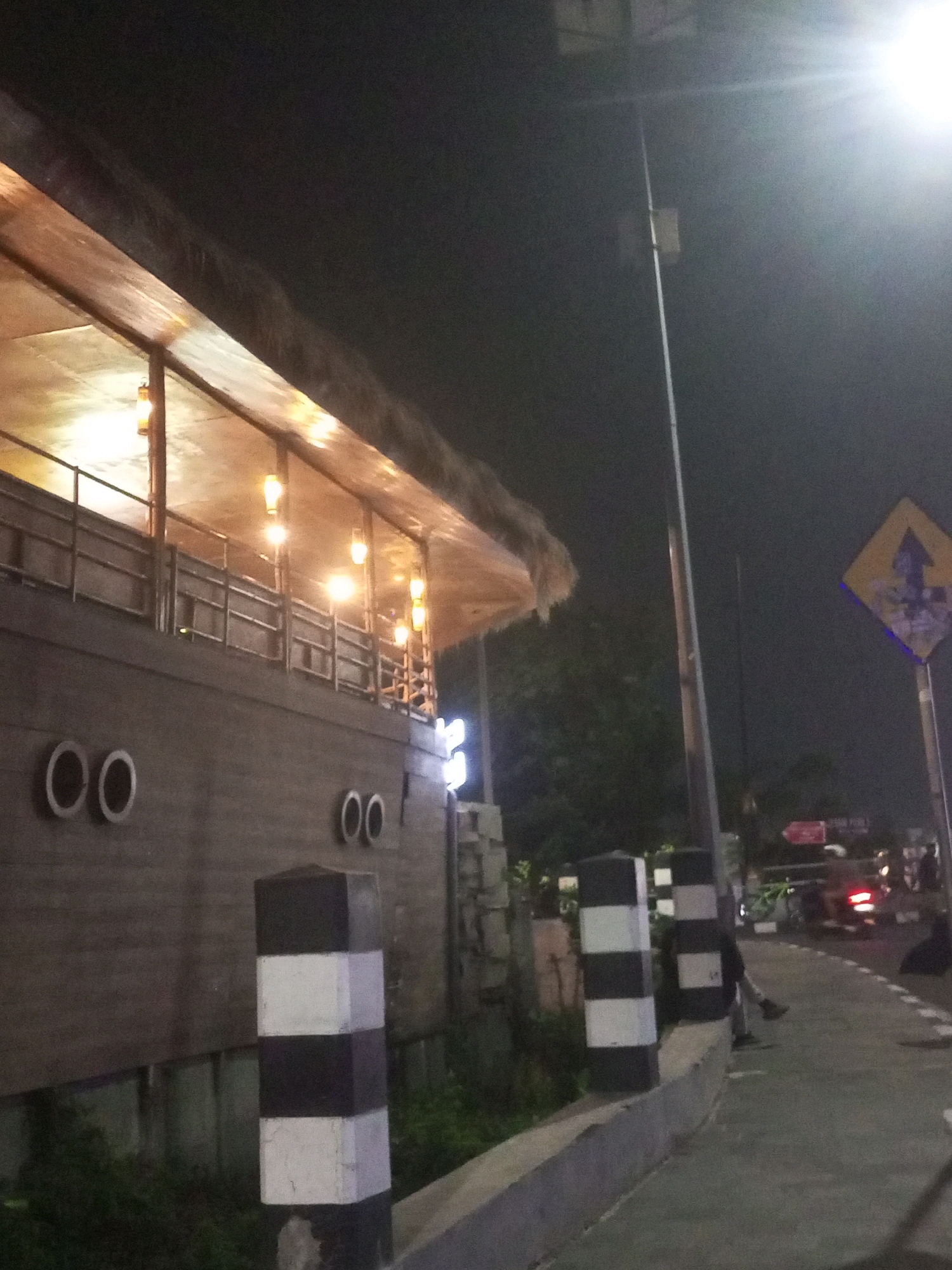 The image depicts a nighttime scene on a street in Kediri City, Indonesia. In the foreground, a concrete barrier lines the left side of the street, topped with two black and white striped bollards. To the left of the bollards, a building with a thatched roof is visible. The building has a wooden exterior, with a railing on an upper level. Several round, dark circular shapes, resembling portholes, are embedded in the lower section of the building. Interior lights are illuminated within the building, casting a warm glow.
The street itself is paved and appears to be an asphalt road with white lane markings. A bright streetlight illuminates the scene from the upper right corner of the image. A yellow diamond-shaped traffic sign with a blue arrow pointing upwards and to the left is mounted on a pole on the right side of the street. Further down the street, several motorcycles and individuals are visible, suggesting a public area or commercial zone. Some of the motorcycles have their headlights and taillights illuminated. The overall environment appears to be urban, with trees and foliage visible in the background. The atmosphere is dimly lit due to the nighttime setting and artificial lighting.