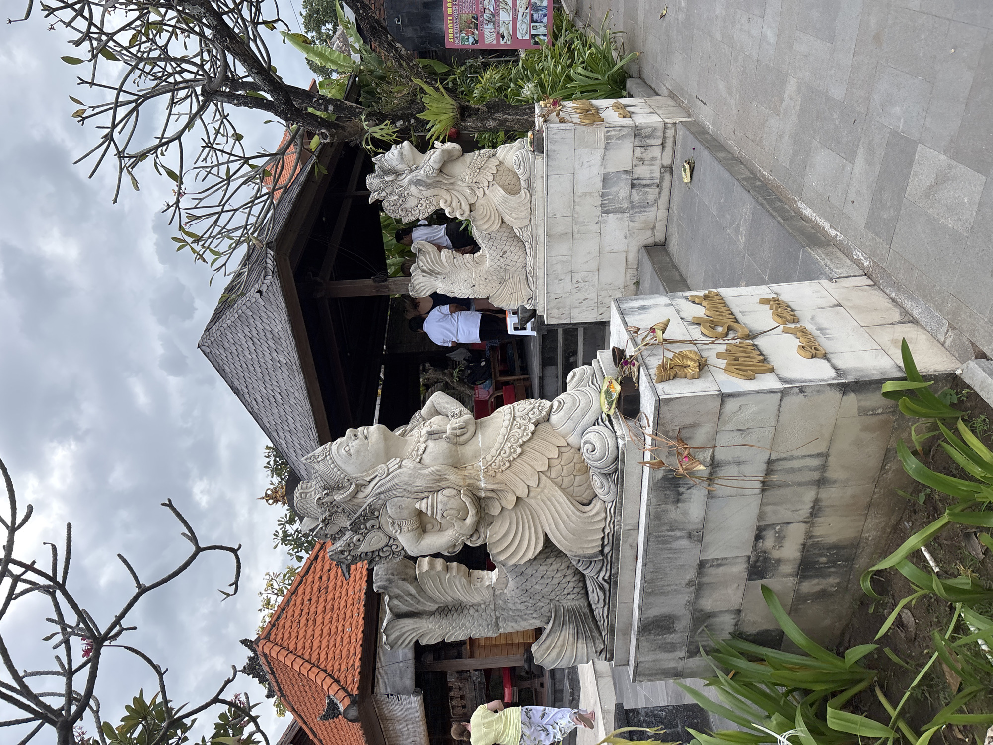 The image shows a detailed view of two traditional Balinese stone sculptures, one in the foreground and another slightly behind it, both depicting mythical creatures with human torsos and fish tails. The sculptures are ornate, with intricate carvings on their headdresses, jewelry, and clothing. They are placed on elevated stone pedestals in what appears to be an outdoor setting, possibly a temple or a spa.
In the background, a traditional Balinese building with a terracotta tiled roof and a wooden pavilion can be seen. Several people are present, some are standing near the pavilion, and one person in a yellow shirt and patterned pants is walking away from the camera on a stone pathway. Lush green foliage, including palm-like plants and trees with bare branches, frames the scene, suggesting a tropical environment. A laminated sign displaying various spa treatments is visible on the right side of the image. The overall ambiance is serene and cultural, typical of tourist destinations in Bali.
This scene is likely located in Denpasar, Bali, Indonesia, given the distinctive architectural style and the presence of Balinese cultural elements like the stone sculptures and offerings. The specific location could be a spa or a tourist attraction that incorporates Balinese art and traditions.