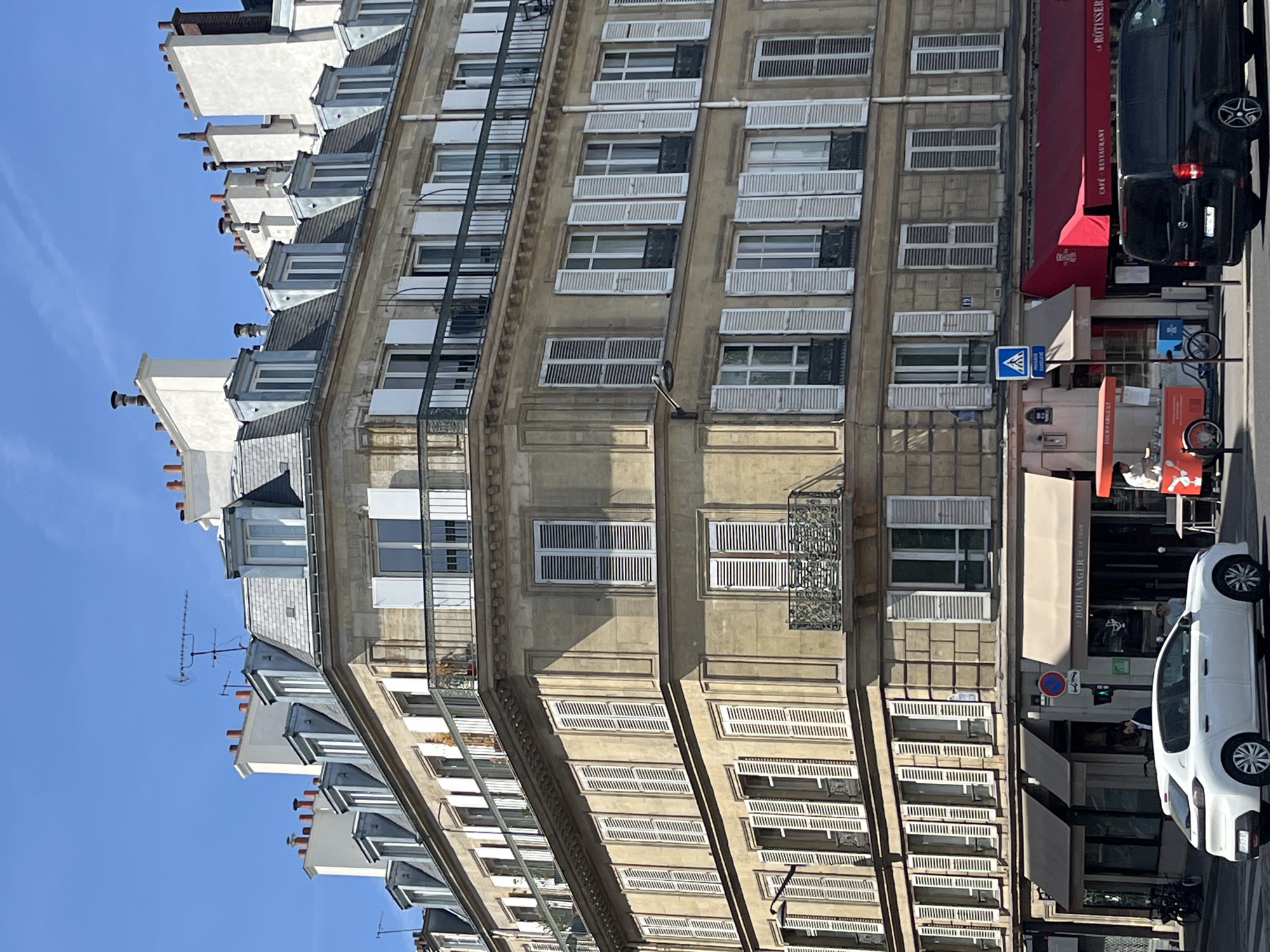 The image captures a corner of a multi-story Haussmannian building in Paris, France, on a sunny day with a clear blue sky. The building features beige stone facades, numerous windows with white shutters, and decorative balconies. The rooftops are adorned with dormer windows and chimneys. At street level, a boulangerie with a beige awning is visible, and next to it, a red awning marks a cafe/restaurant. A white Peugeot 208 is parked in the foreground on the left, and a black Mercedes-Benz Vito van is in the foreground on the right. Pedestrians are present, including someone standing at a street vendor cart and another near the boulangerie entrance. Traffic signals are visible, indicating an intersection. The overall scene depicts a typical Parisian streetscape with architectural charm and daily urban activity.